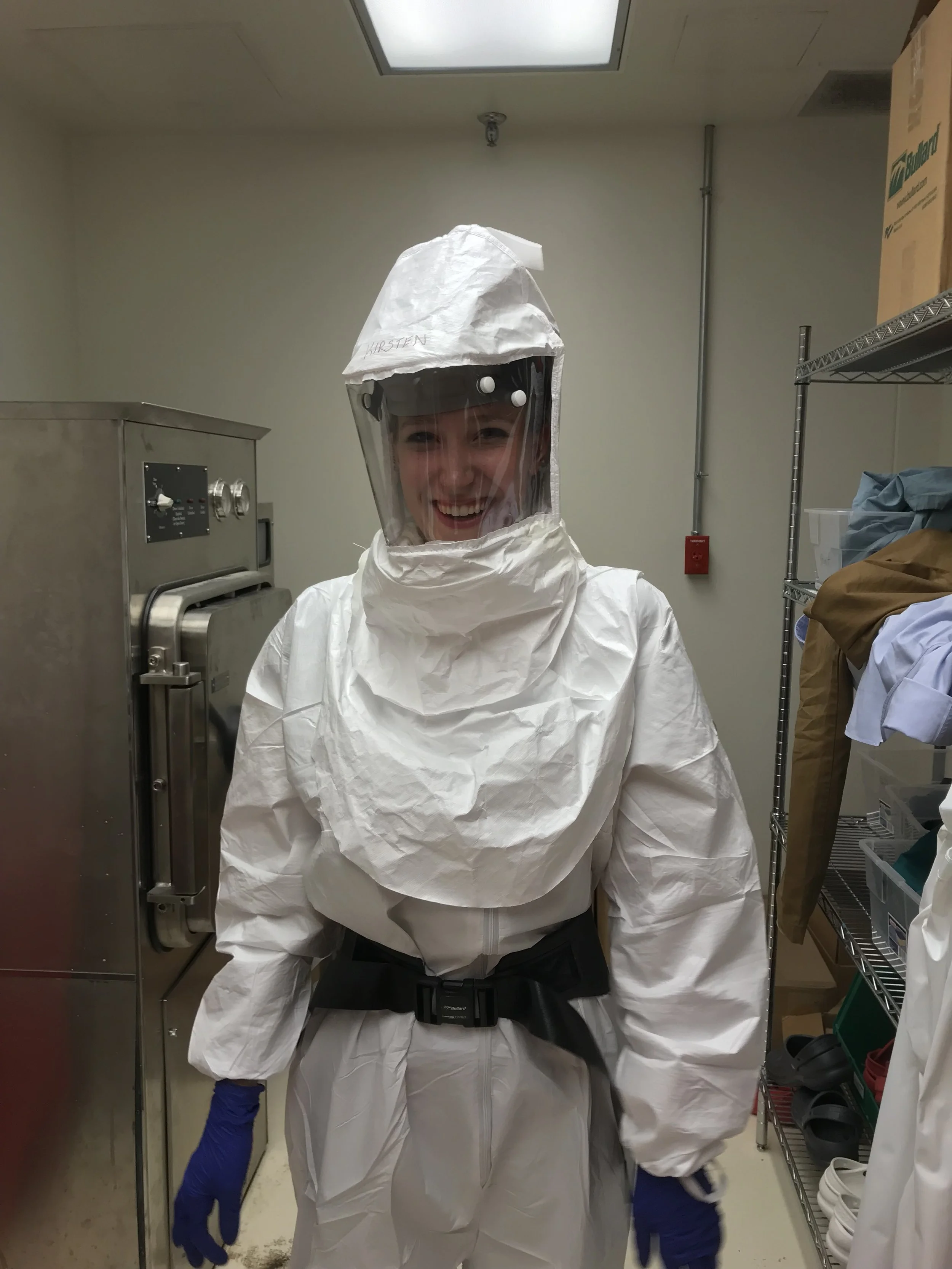 A person wearing full protective biohazard suit, including helmet with face shield, gloves, and a black belt, smiling in a laboratory or cleanroom setting.