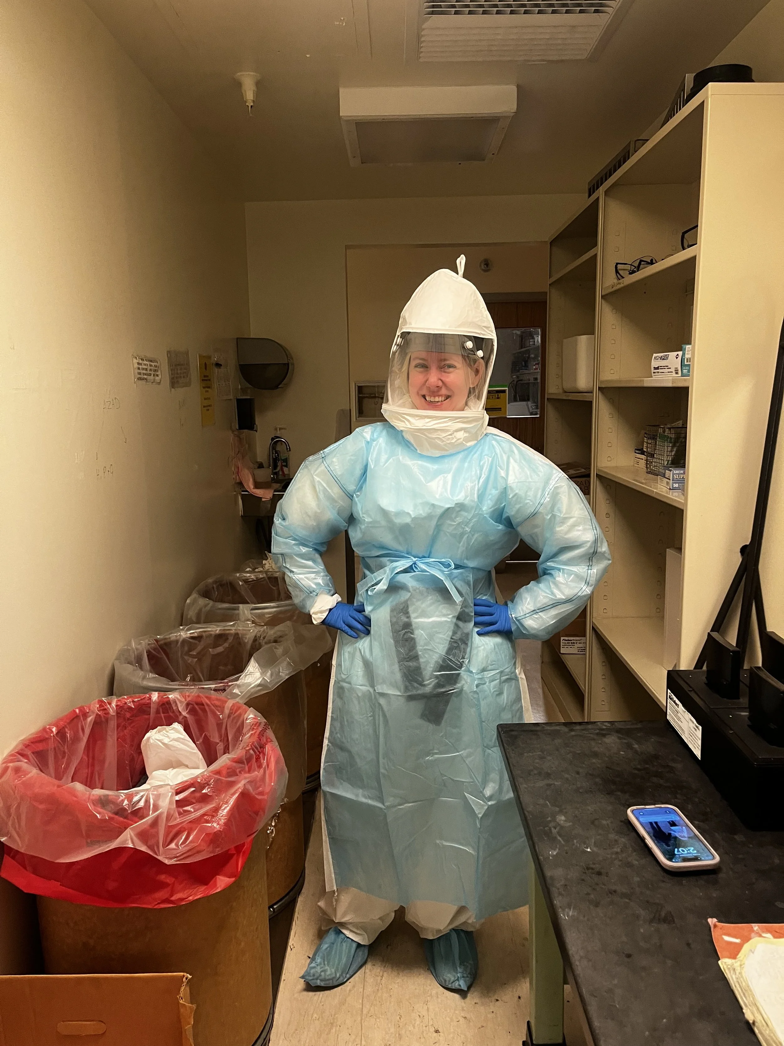 Healthcare worker in full personal protective equipment (PPE), including a gown, gloves, face shield, and hood, standing confident in a medical or laboratory storage room.