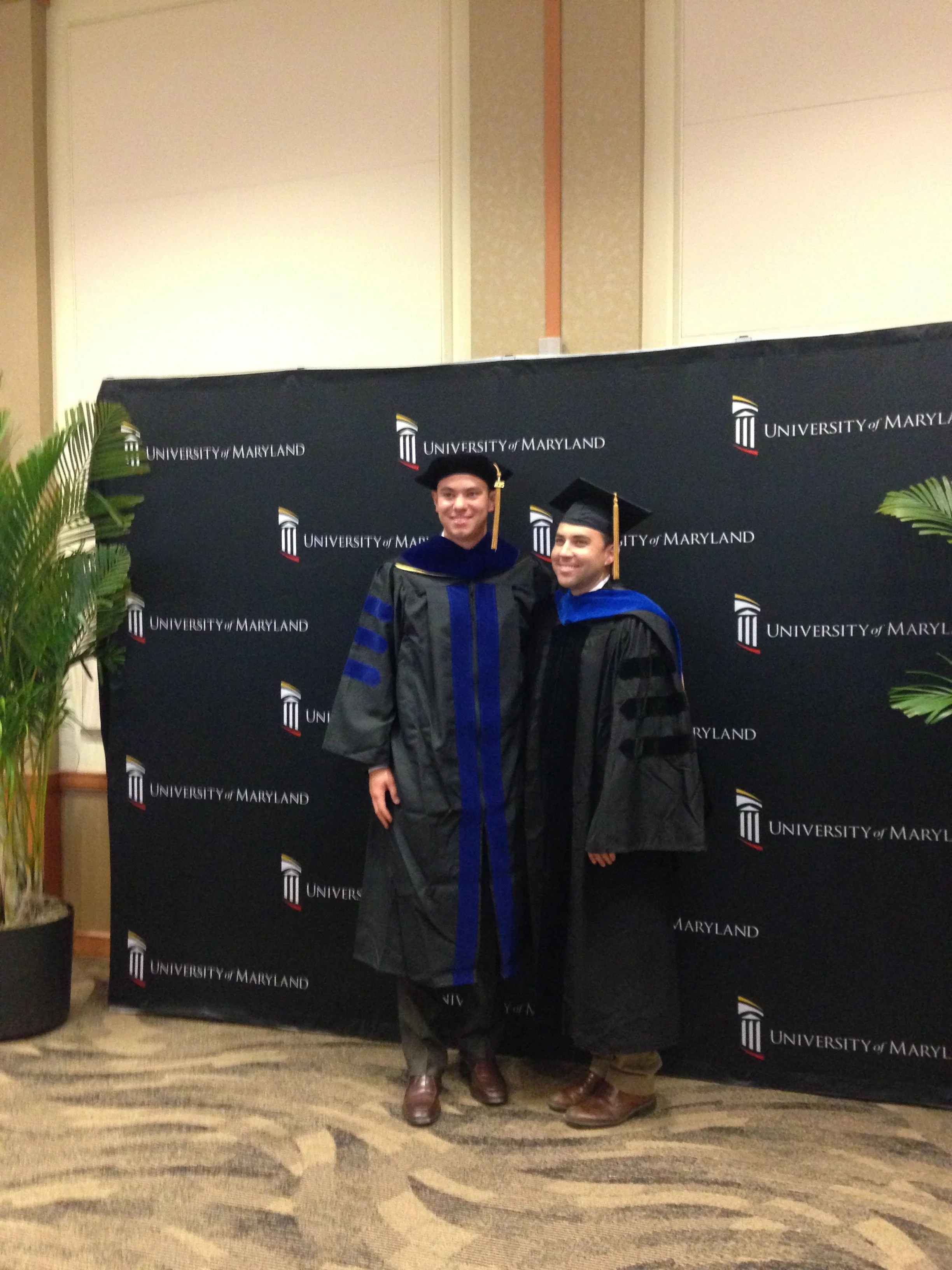 Two men in graduation gowns and caps standing next to each other in front of a backdrop with the University of Maryland logo, smiling for a photo.