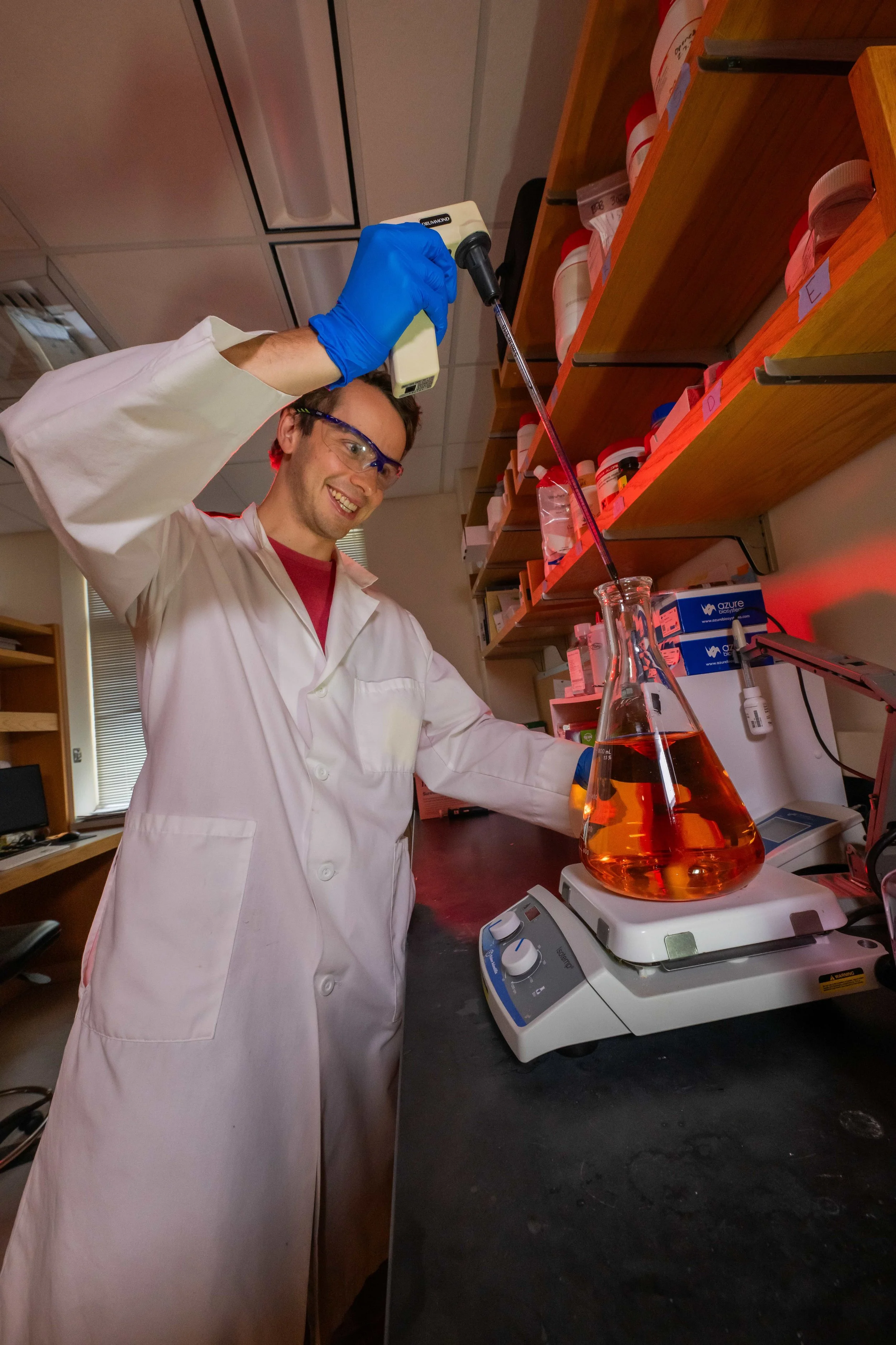 A scientist wearing safety glasses, blue gloves, and a white lab coat is pipetting a red liquid into a conical flask on a magnetic stirrer in a laboratory.