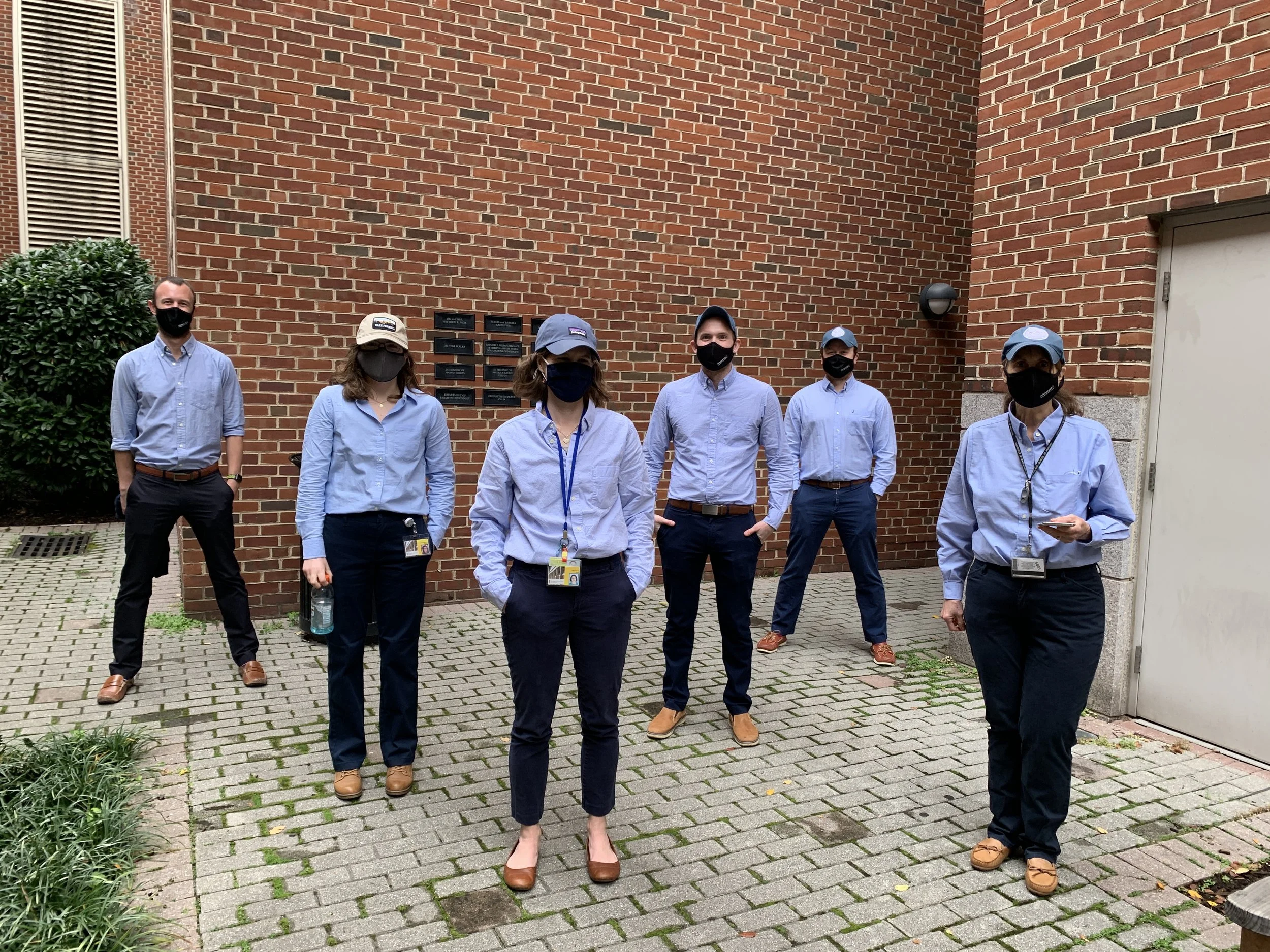Group of six people standing outdoors in front of a brick building, all wearing masks, light blue shirts, and name badges.