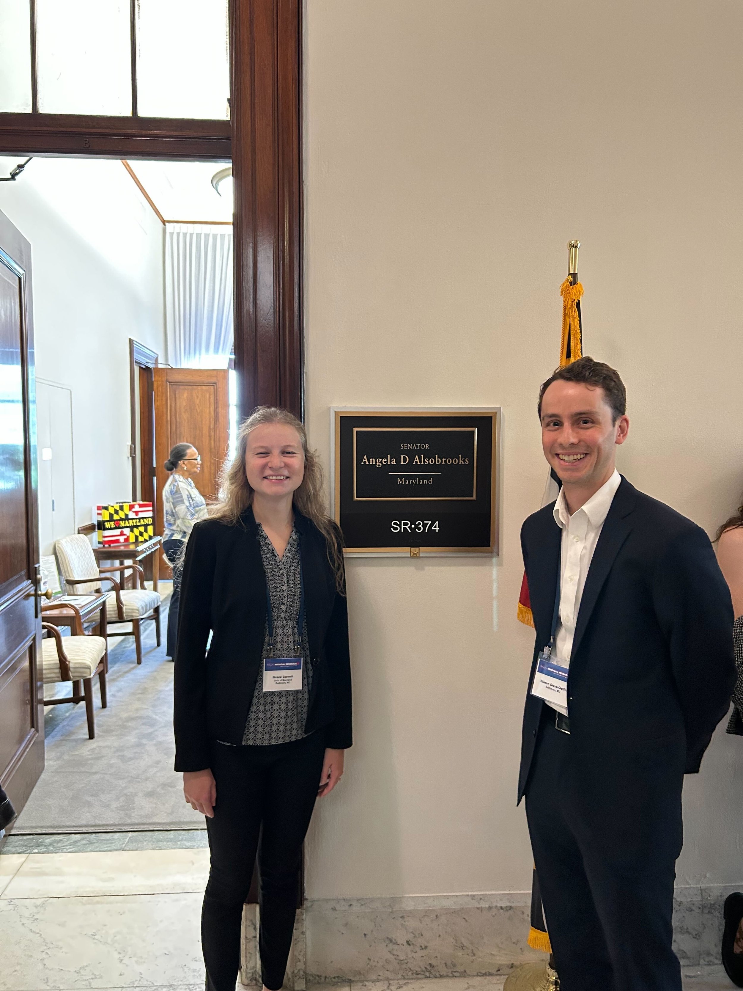 Two individuals in formal attire standing next to a sign that reads "Senator Angela D Alsobrooks Maryland SR-374" inside a building. The woman on the left has long blonde hair, and the man on the right has short dark hair. Both are smiling.