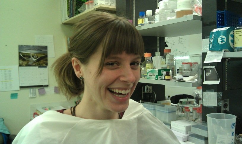 A young woman in a laboratory wearing a white lab coat, smiling, with shelves of lab supplies and equipment in the background.