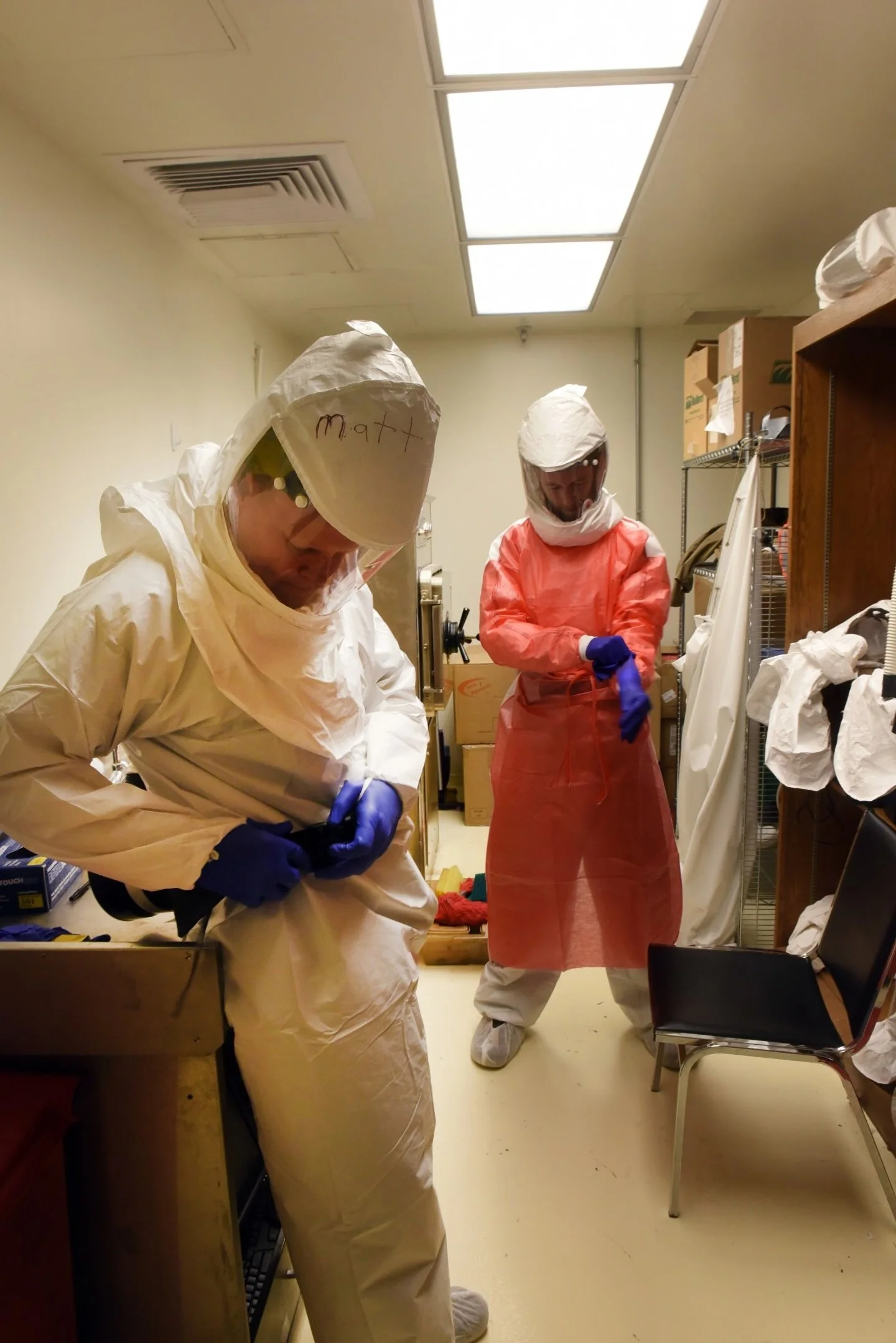 Two people wearing full protective hazmat suits and purple gloves, working in a laboratory or medical setting, with shelves, boxes, and equipment around them.