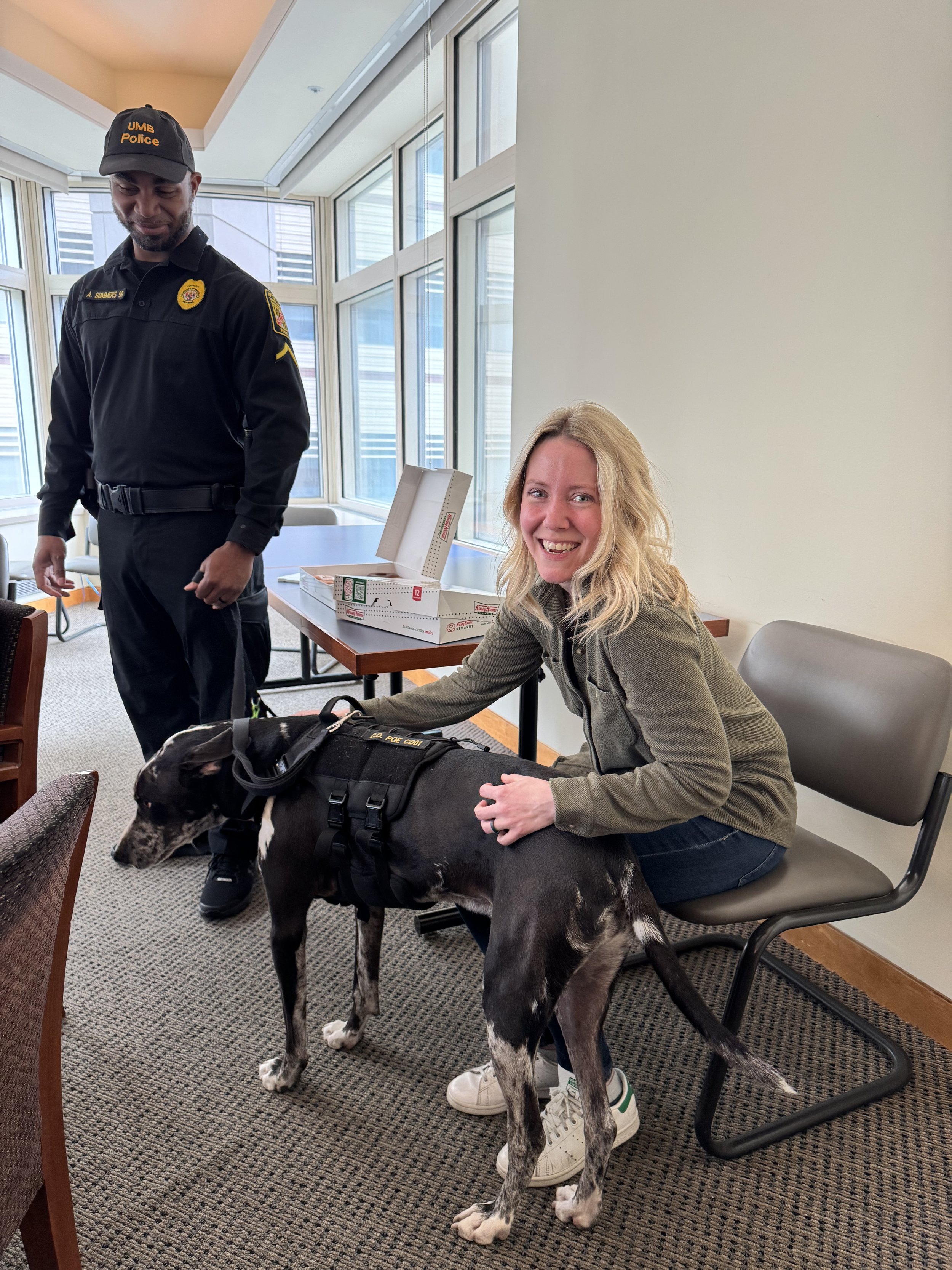 A woman smiling while petting a greyhound dog in a room with large windows, with a police officer standing nearby.
