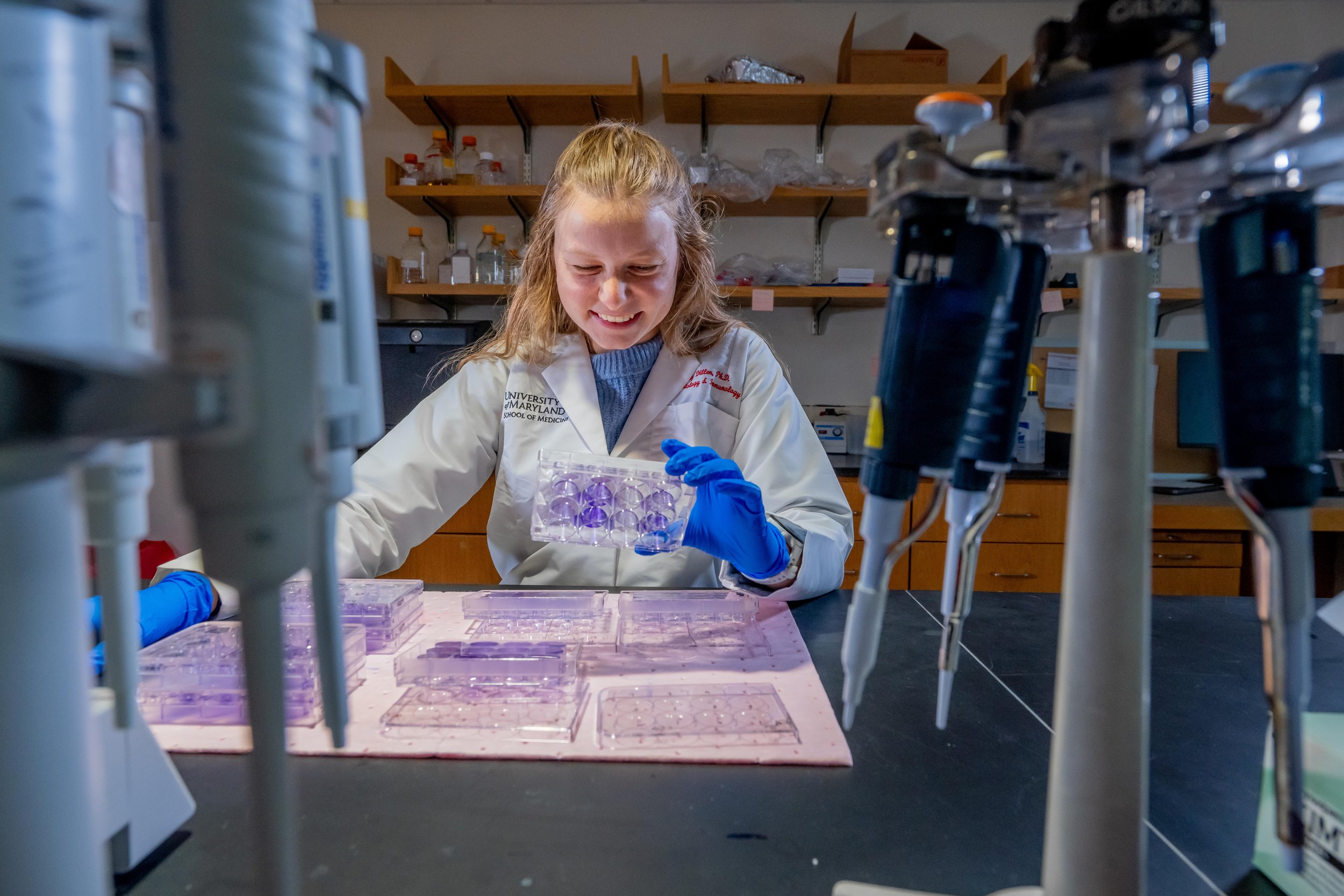 A young woman scientist in a lab coat and blue gloves holding a tray of purple gel electrophoresis wells, working in a laboratory with scientific equipment and supplies.