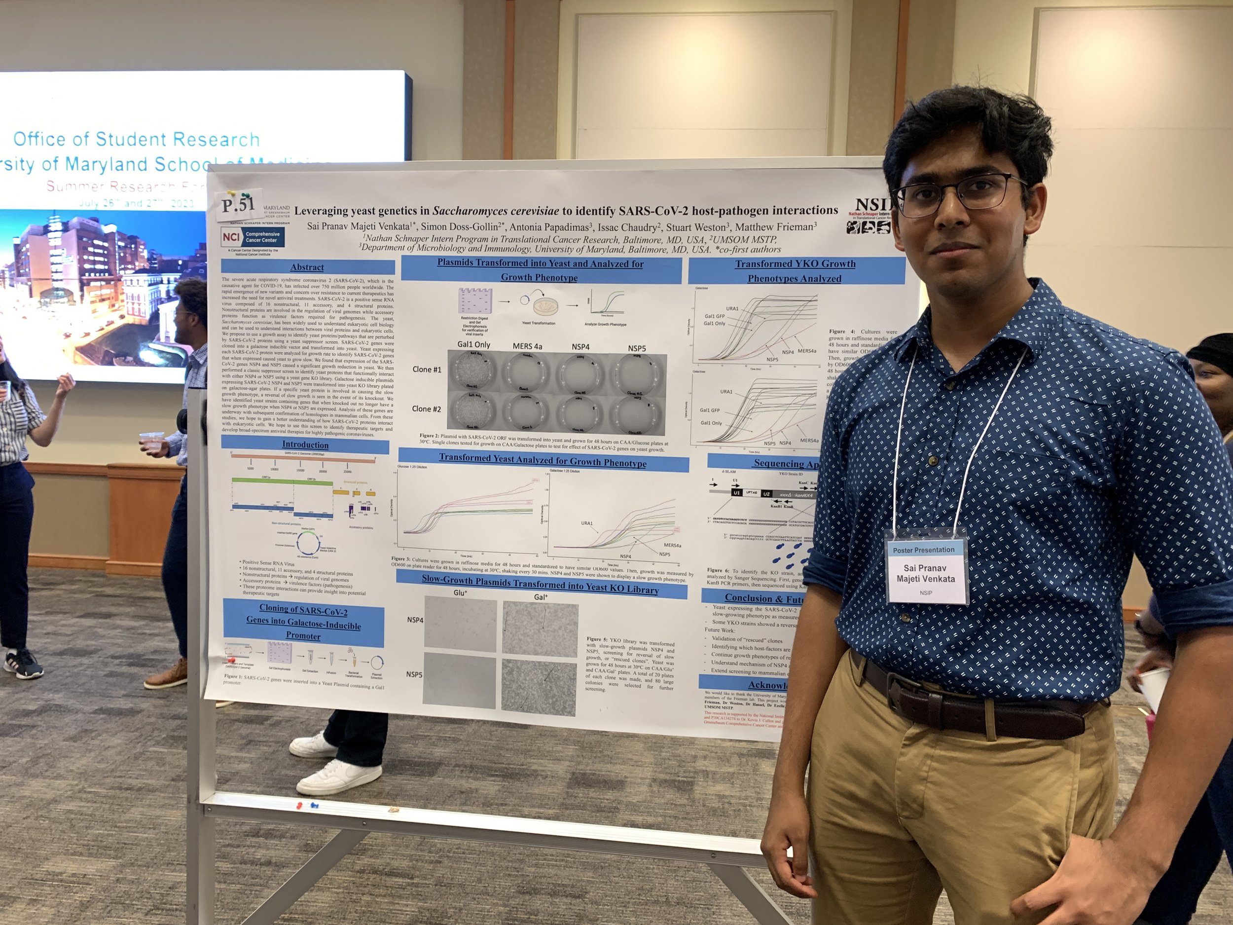 Young man in glasses and blue patterned shirt standing next to a scientific research poster at a conference. The poster is about yeast genetics and SARS-CoV-2 interactions.