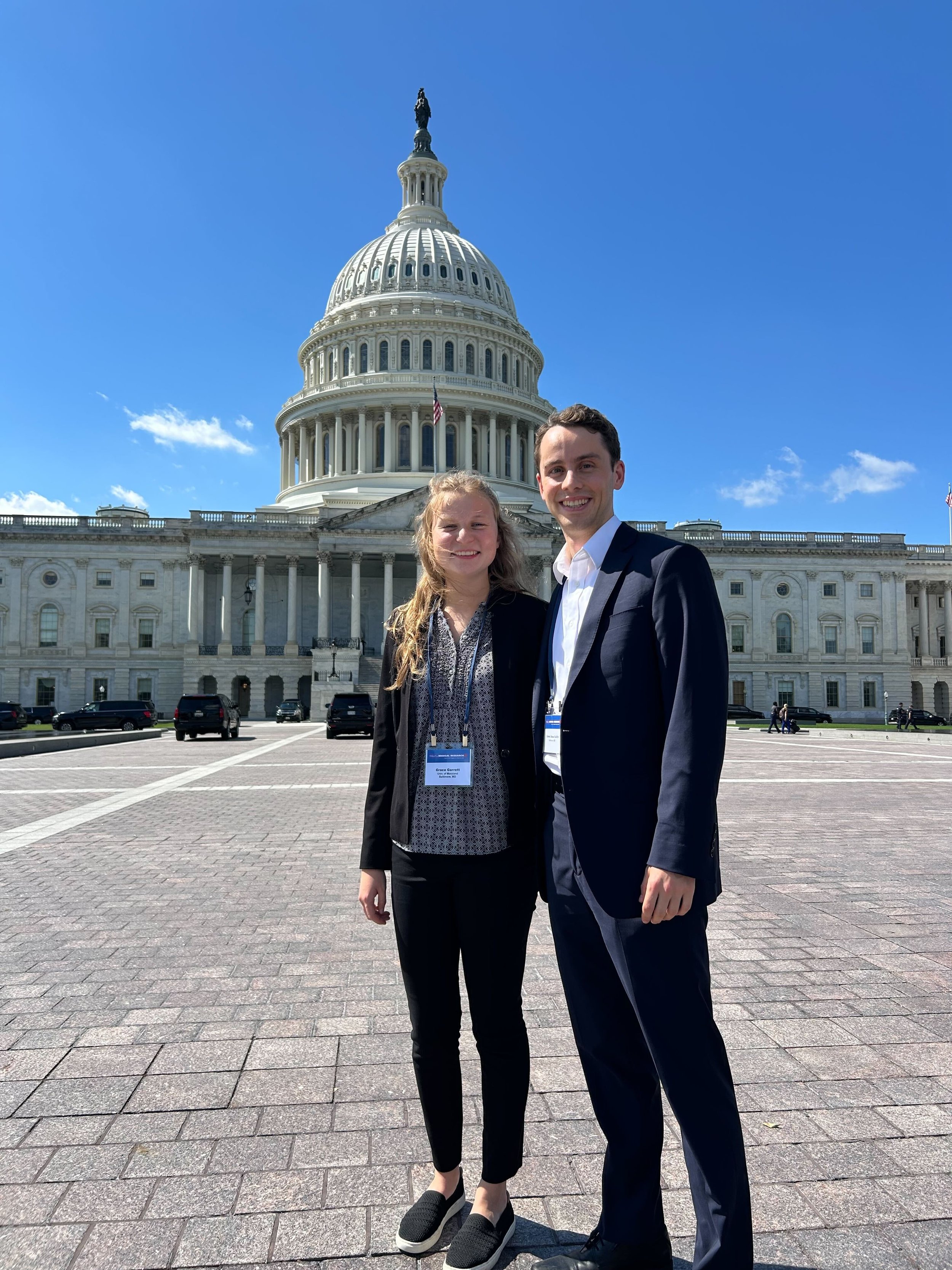 A woman and a man in business attire standing outside in front of the U.S. Capitol building on a sunny day, smiling at the camera.