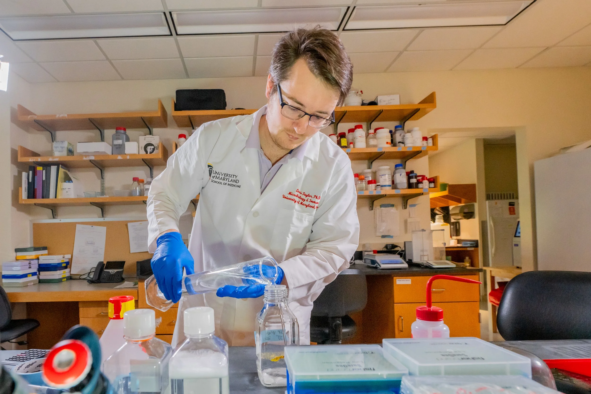 A man wearing a white lab coat and blue gloves working in a laboratory, pouring liquid from a beaker into a smaller container. Shelves with bottles and office supplies are visible behind him.