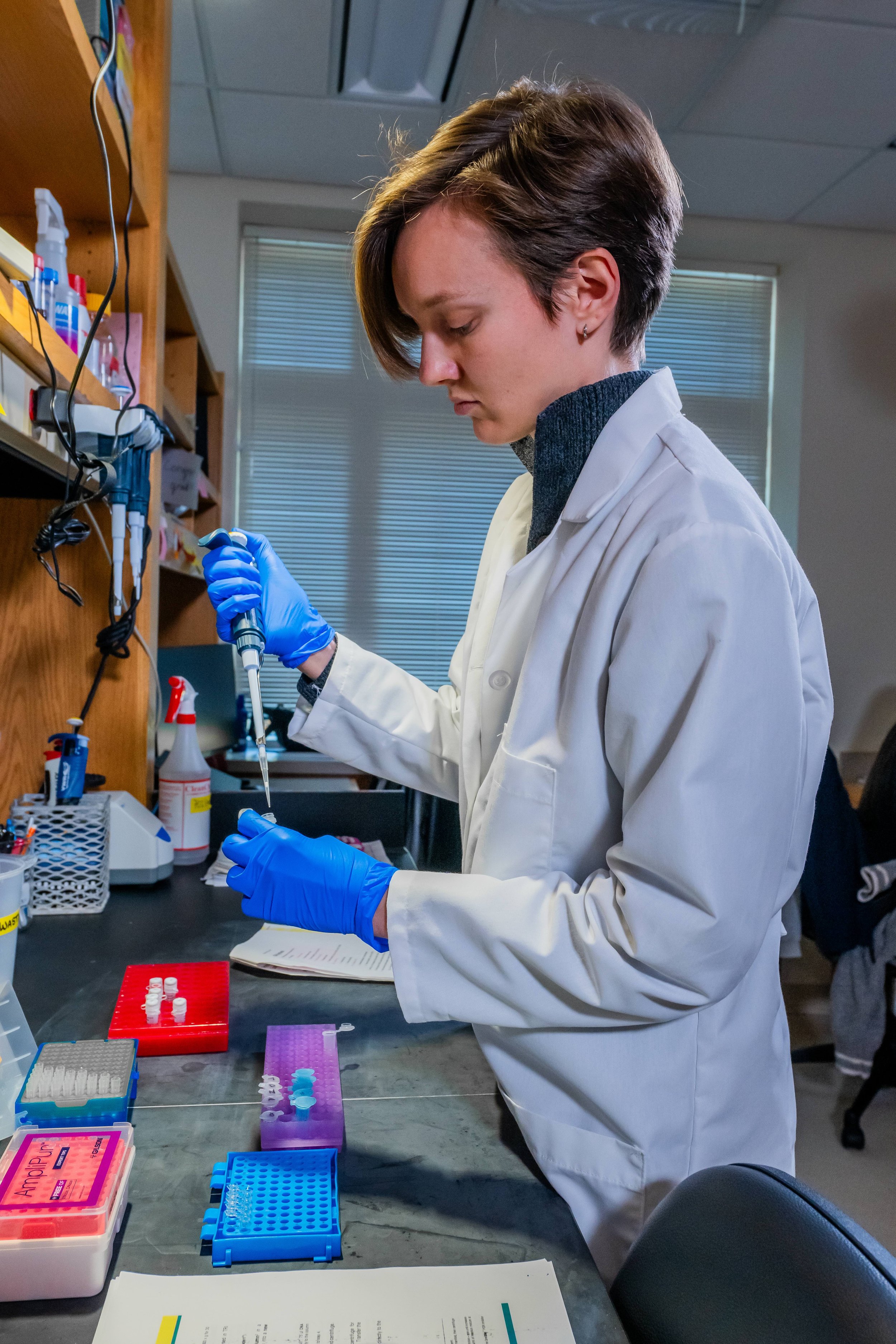A scientist with short brown hair working in a laboratory, wearing a white lab coat and blue gloves, using a pipette to transfer liquid. The lab has various scientific equipment and supplies on the counter.