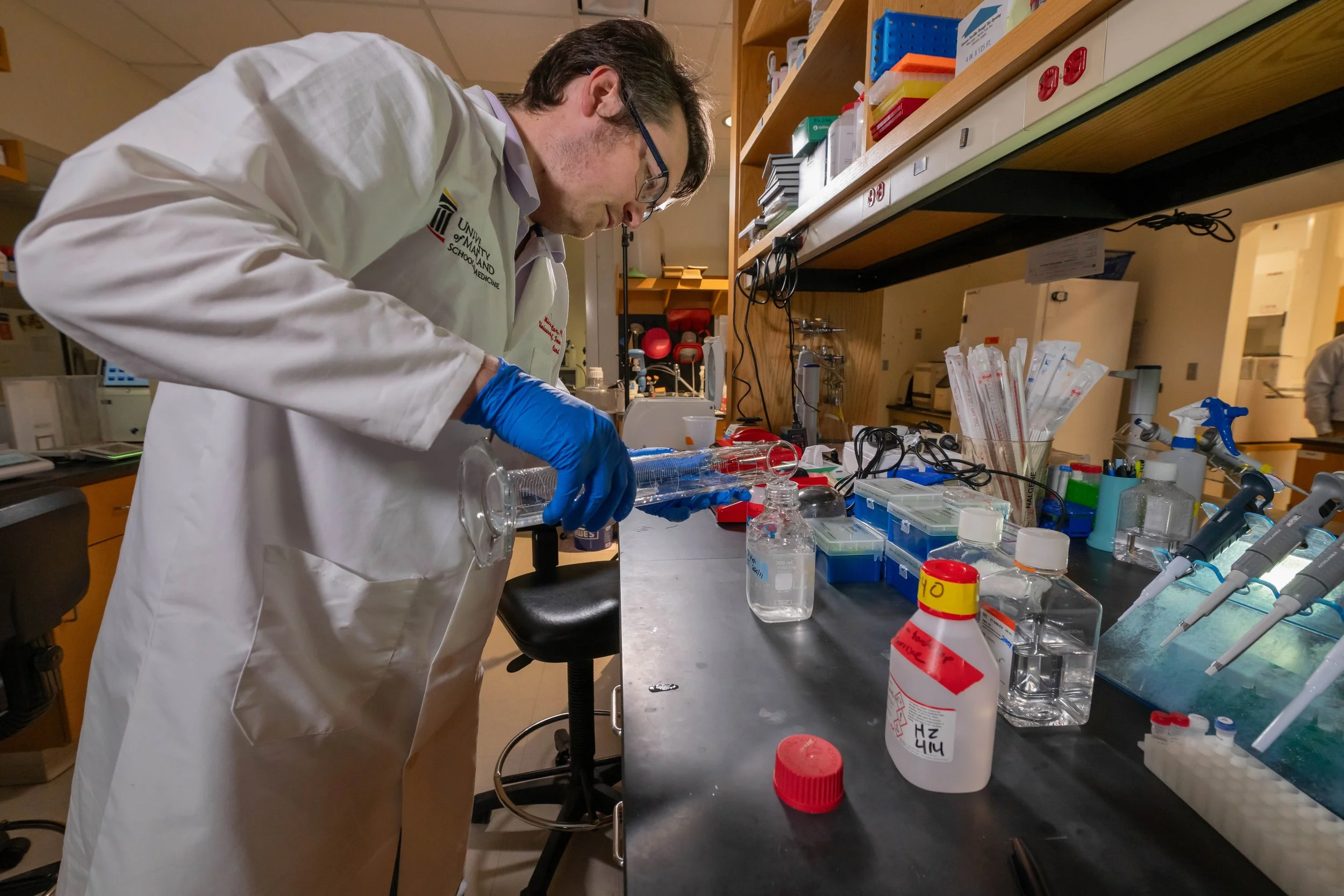 A scientist in a lab coat and blue gloves working with laboratory equipment on a black workbench in a lab.