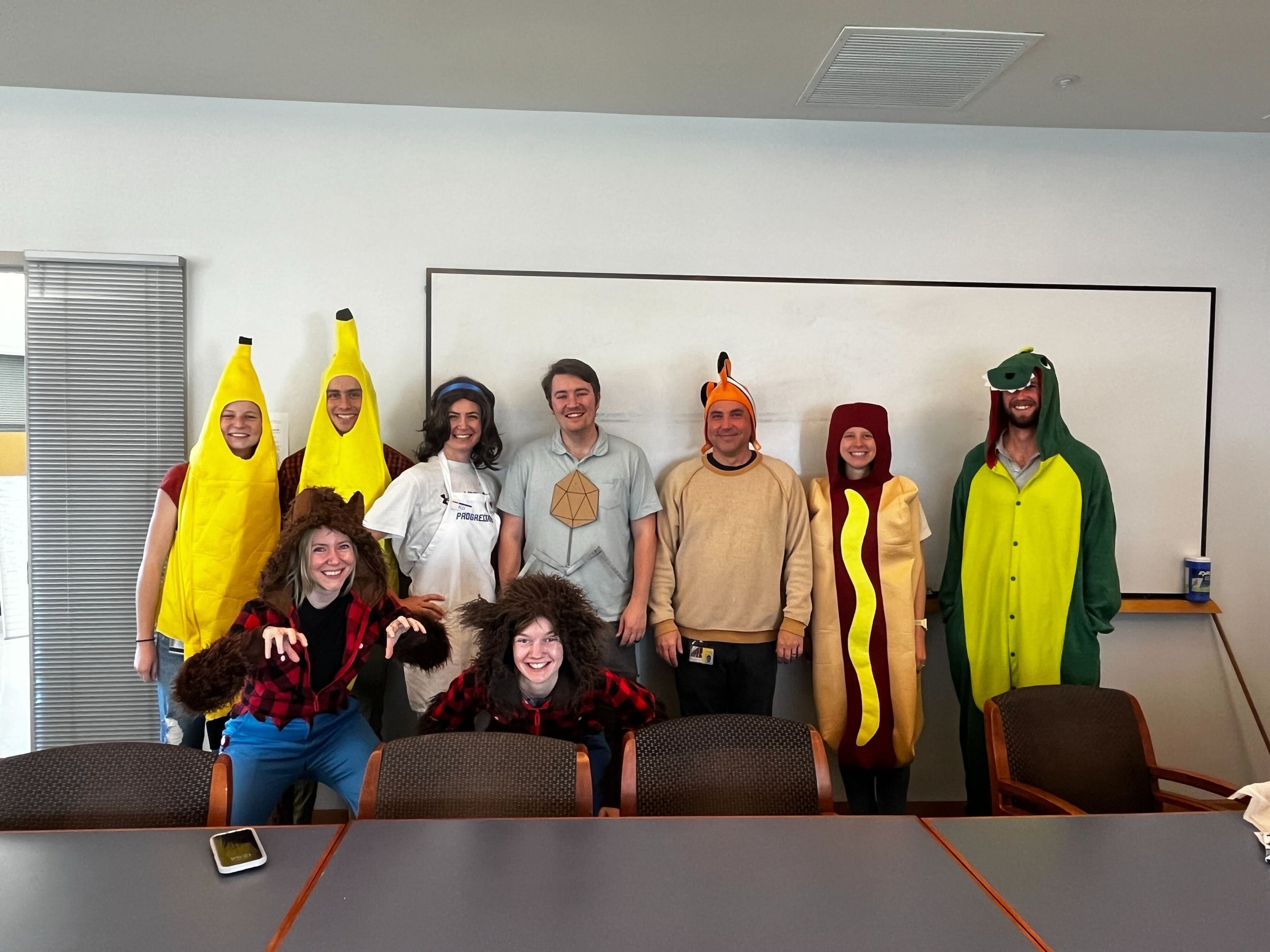 Group of nine people in fruit and animal costumes, smiling and posing in an office conference room.