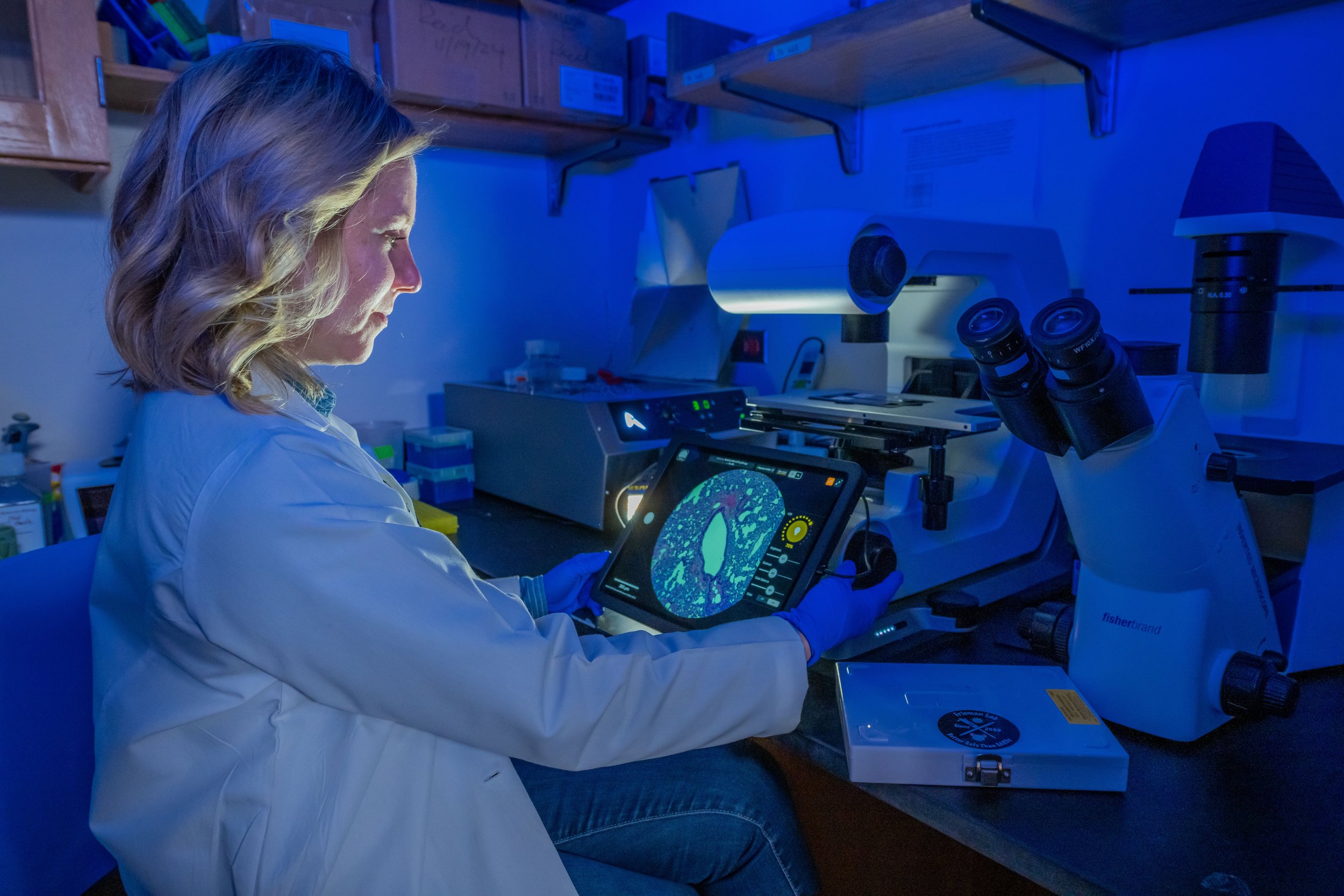 A female scientist in a laboratory examining samples through a computer tablet with a digital interface, using a microscope and surrounded by laboratory equipment in a blue-lit setting.