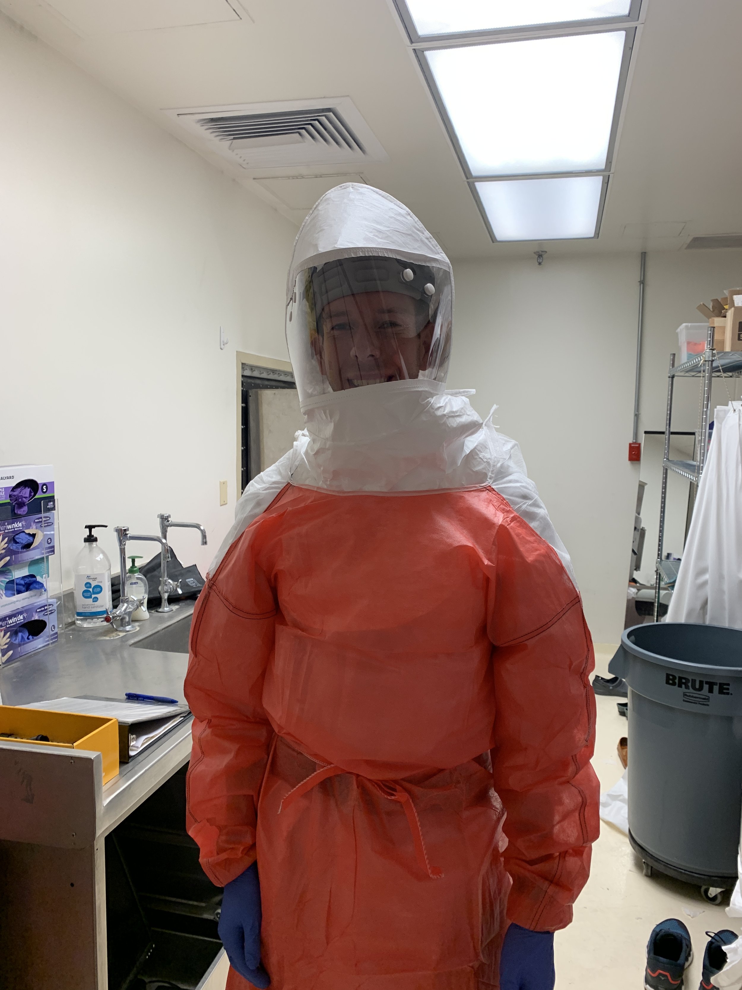 Person wearing full hazmat protective suit, including face shield, in a medical or laboratory setting.