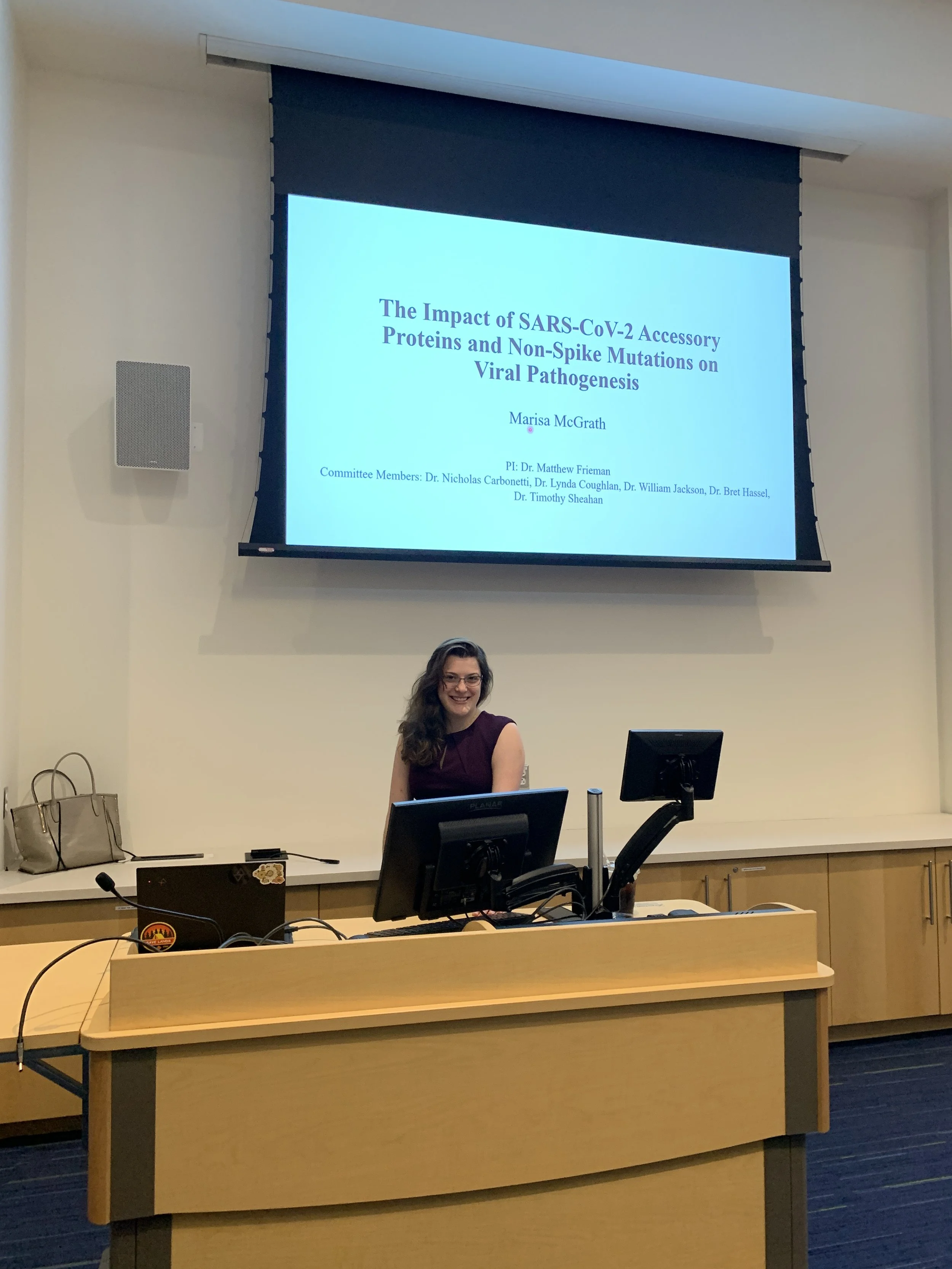 A woman standing behind a lecture podium with computer monitors, smiling at the camera, in a classroom or conference room with a presentation slide projected behind her.