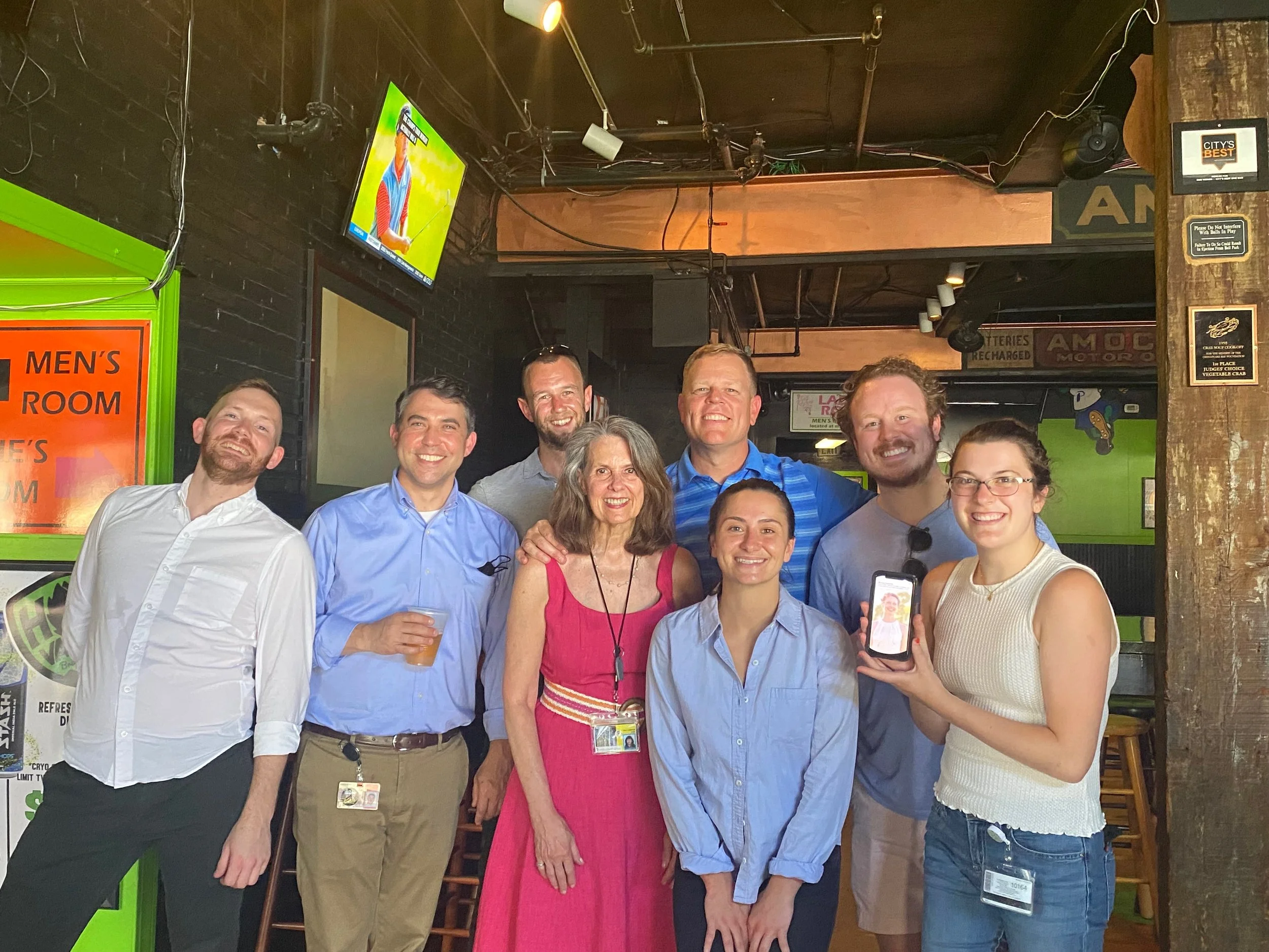 Group of nine smiling people standing in a bar, some wearing glasses, three holding drinks, and one person showing a phone with a picture on it. There are television screens and various signs in the background.