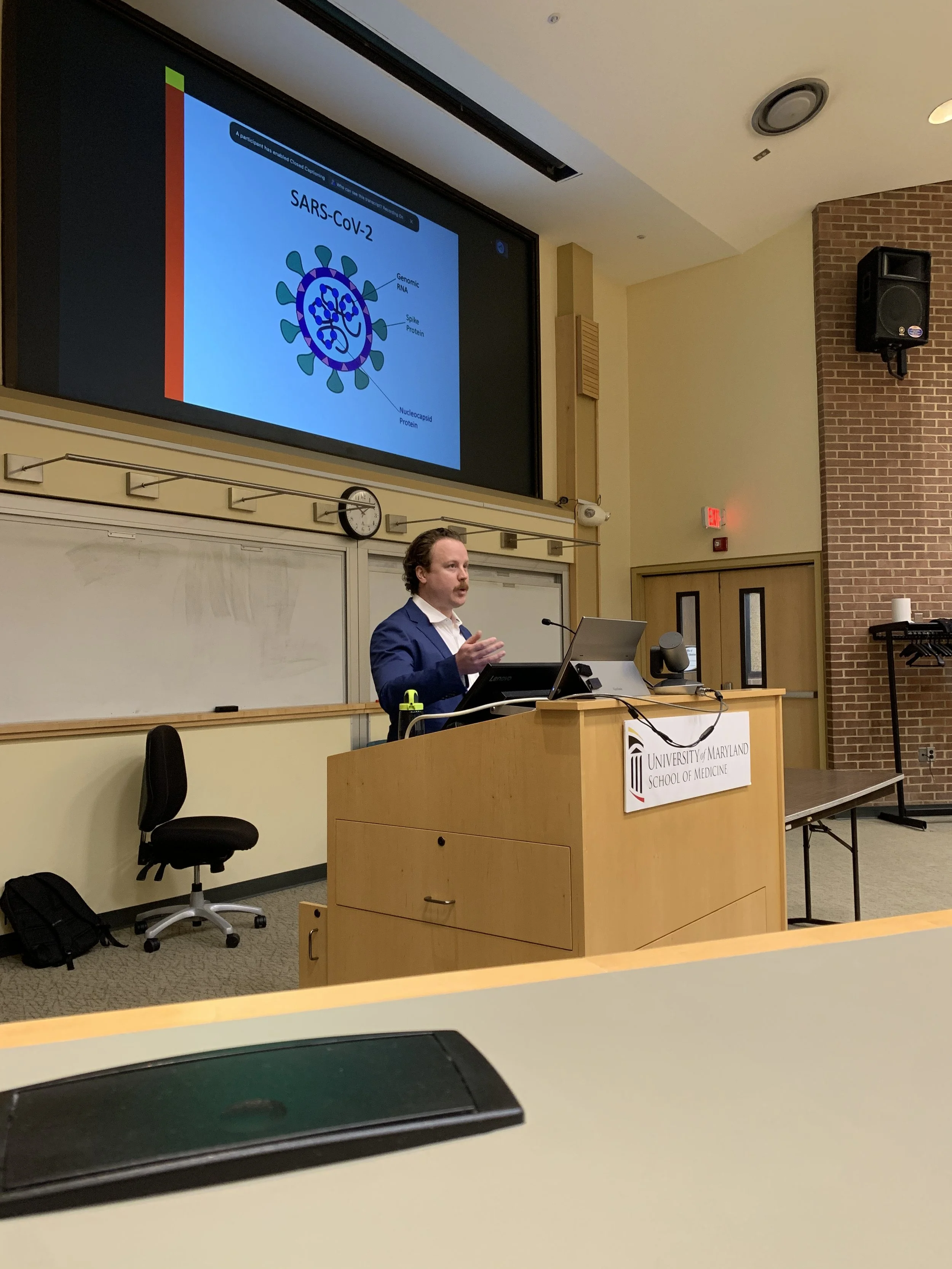 A man in a blue suit delivering a lecture at the University of Maryland School of Medicine, with a presentation slide on SARS-CoV-2 projected behind him.