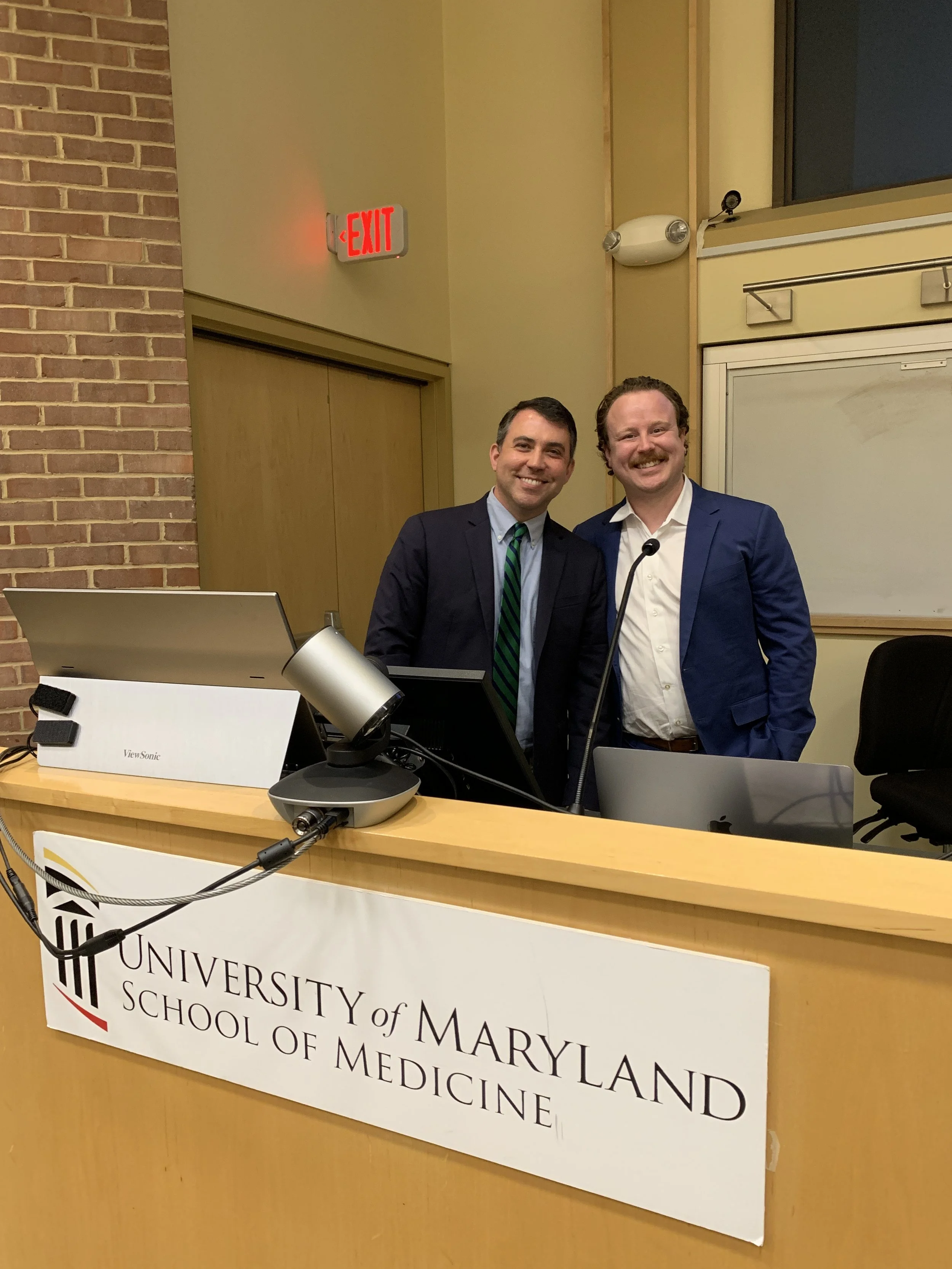 Two men in suits smiling at a university event at the University of Maryland School of Medicine, standing behind a podium with a sign, in a room with brick and yellow walls and an exit sign.