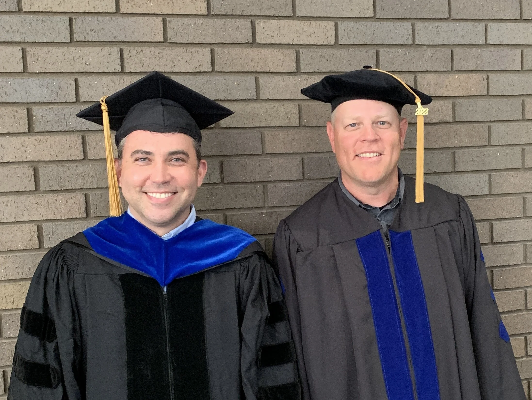 Two men in academic regalia standing in front of a brick wall, both wearing black graduation caps with tassels. The man on the left has dark hair and a big smile, wearing a black gown with a blue velvet hood. The man on the right has light hair, a sl