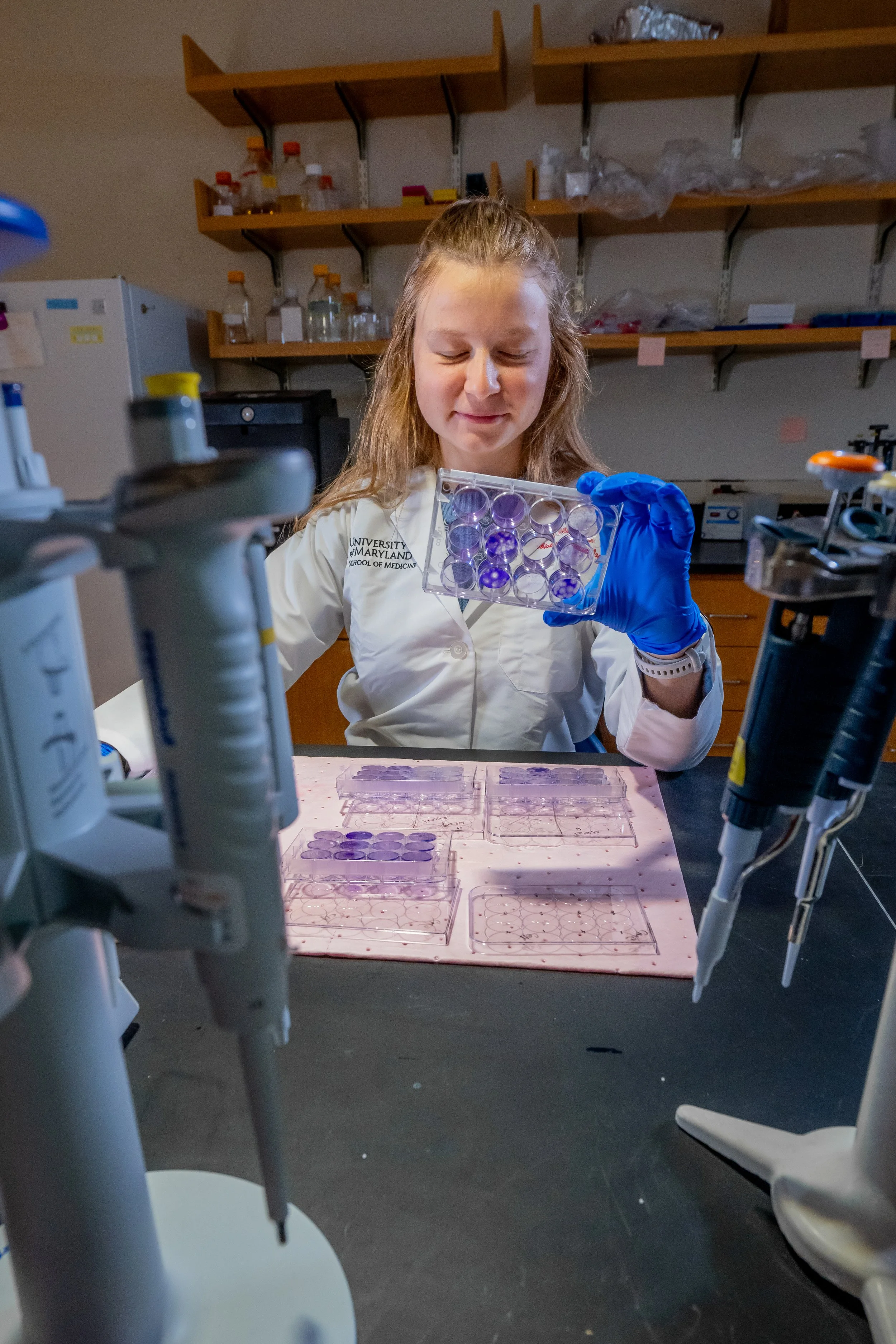 A young woman in a lab coat holding a tray of purple test tubes, working in a science laboratory with equipment and shelves in the background.