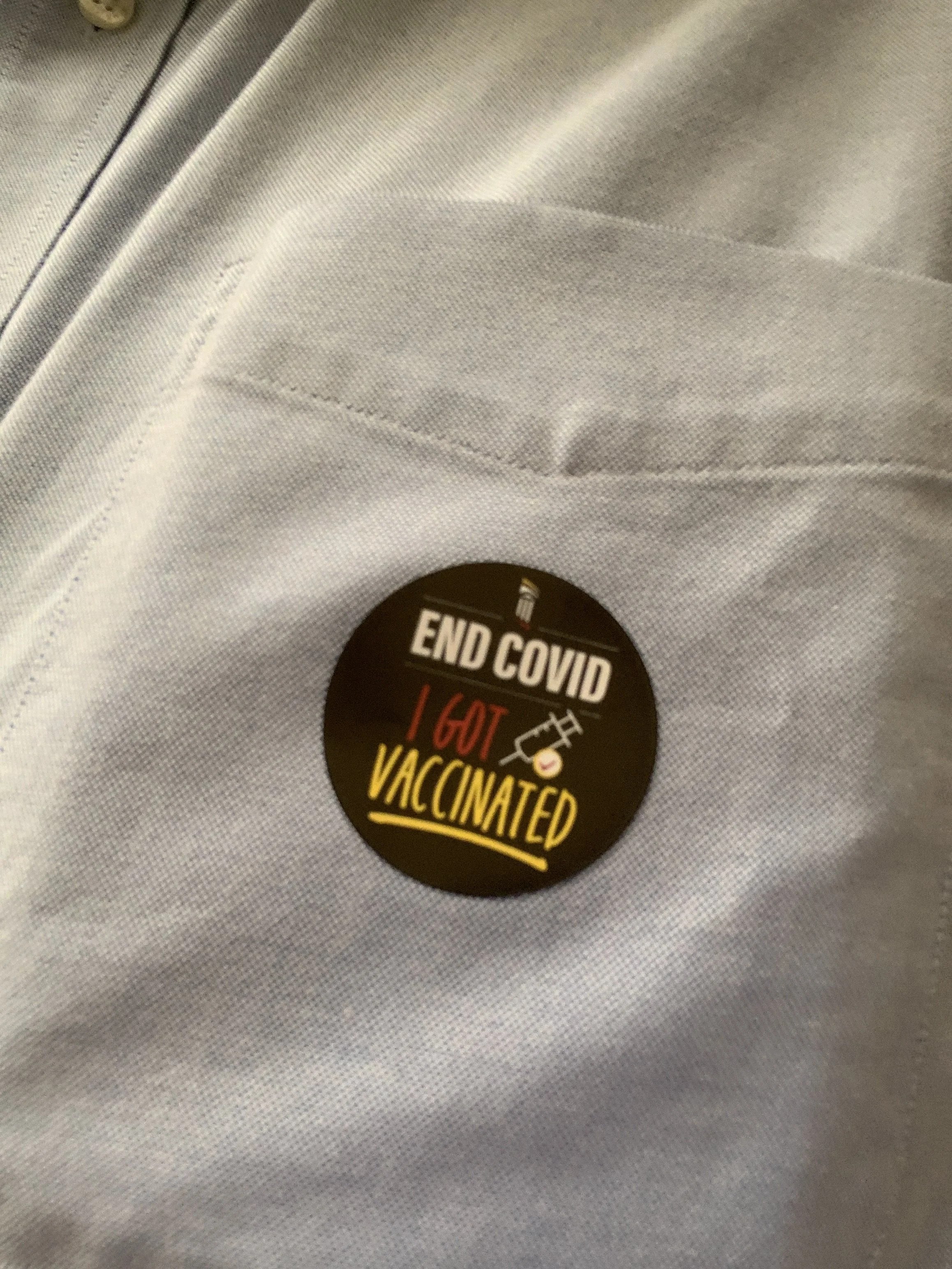 A close-up photo of a round black sticker on light-colored fabric with the text 'END COVID I GOT VACCINATED' and a small graphic of a syringe.