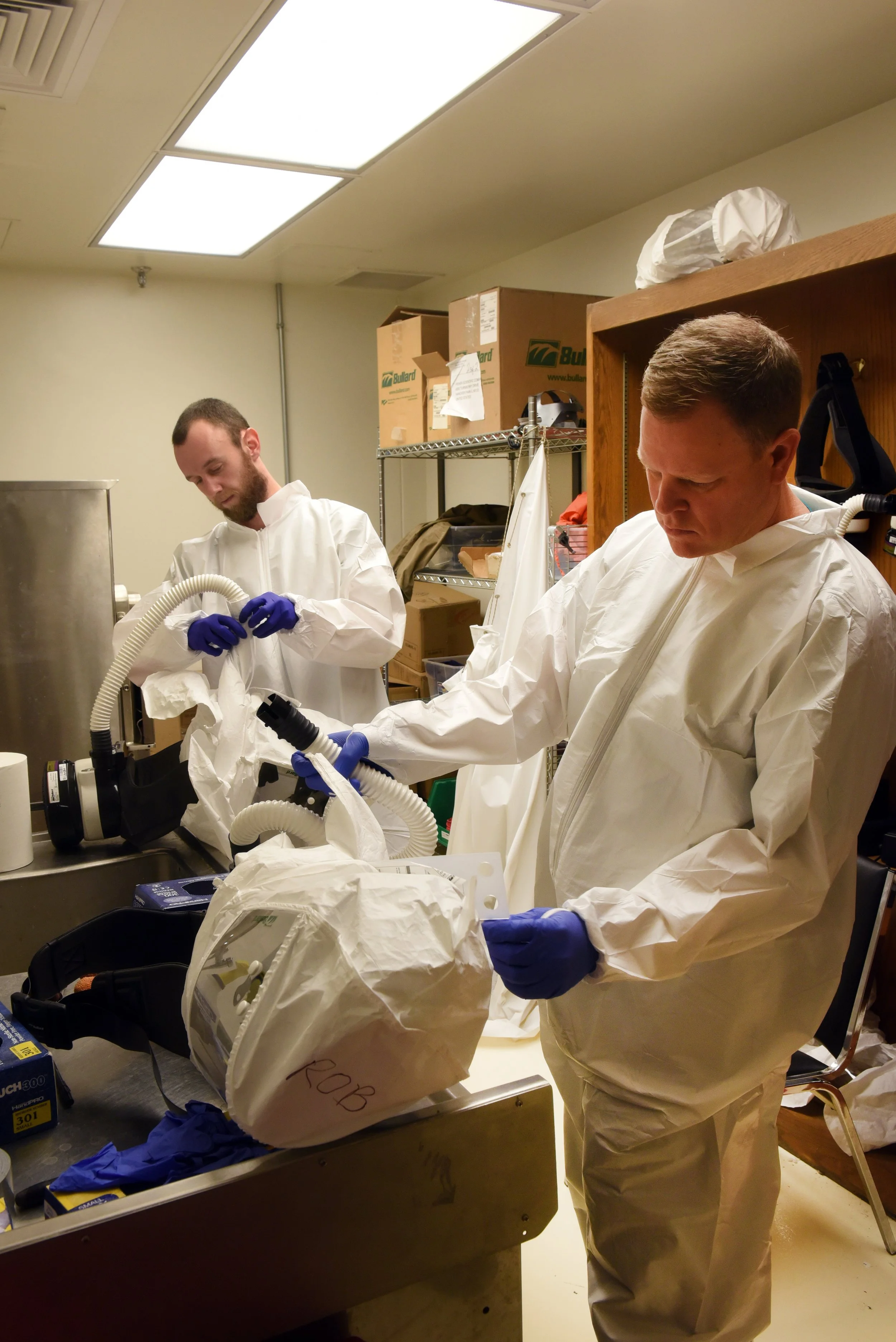 Two researchers wearing white protective suits and blue gloves working with medical equipment and ventilators in a clinical setting.