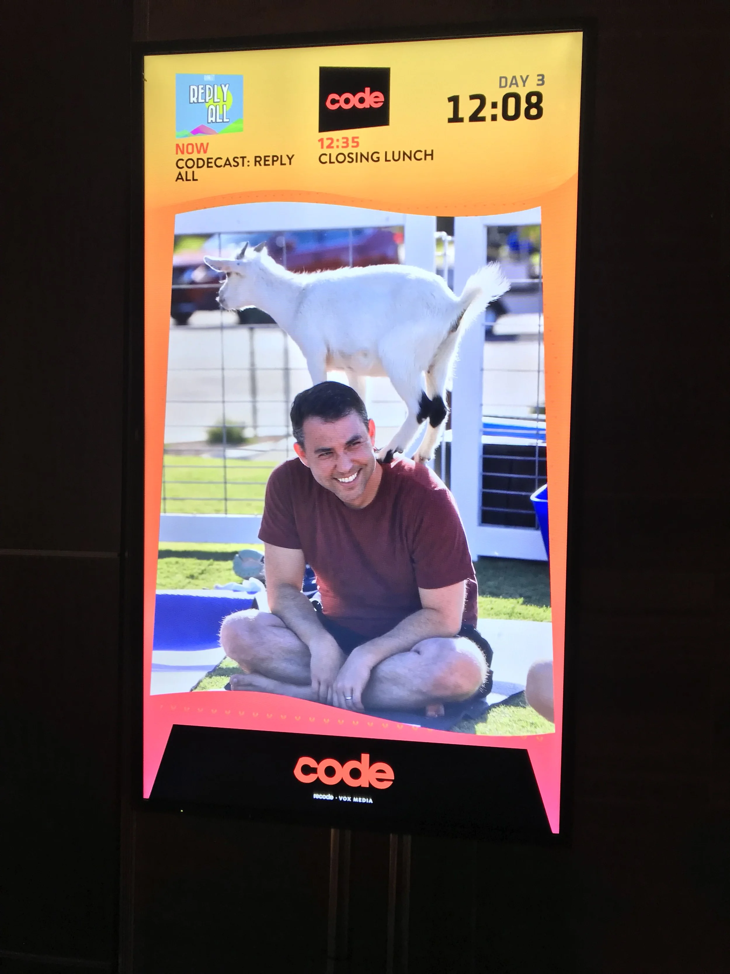 Digital display board showing schedule and a photo of a man sitting cross-legged on the grass with a goat standing on his shoulders, smiling.