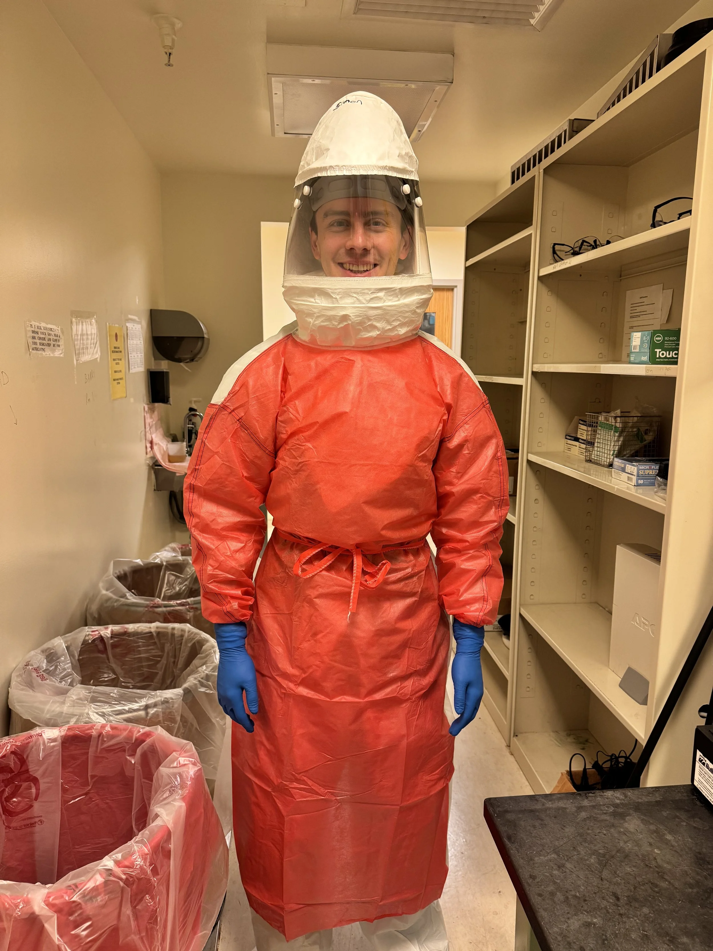 Person wearing a full hazmat suit, including a red coverall, gloves, and a protective helmet with a face shield, standing in a room with shelves and trash bins.