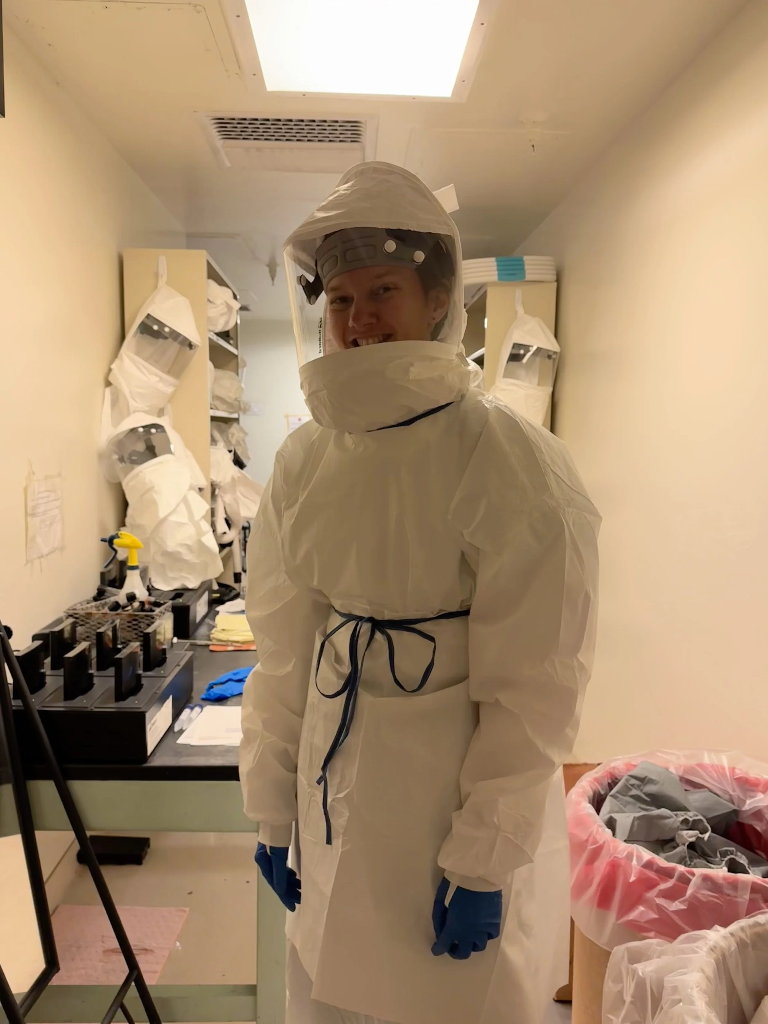 Person wearing full protective hazmat suit and face shield inside a hospital or laboratory room.