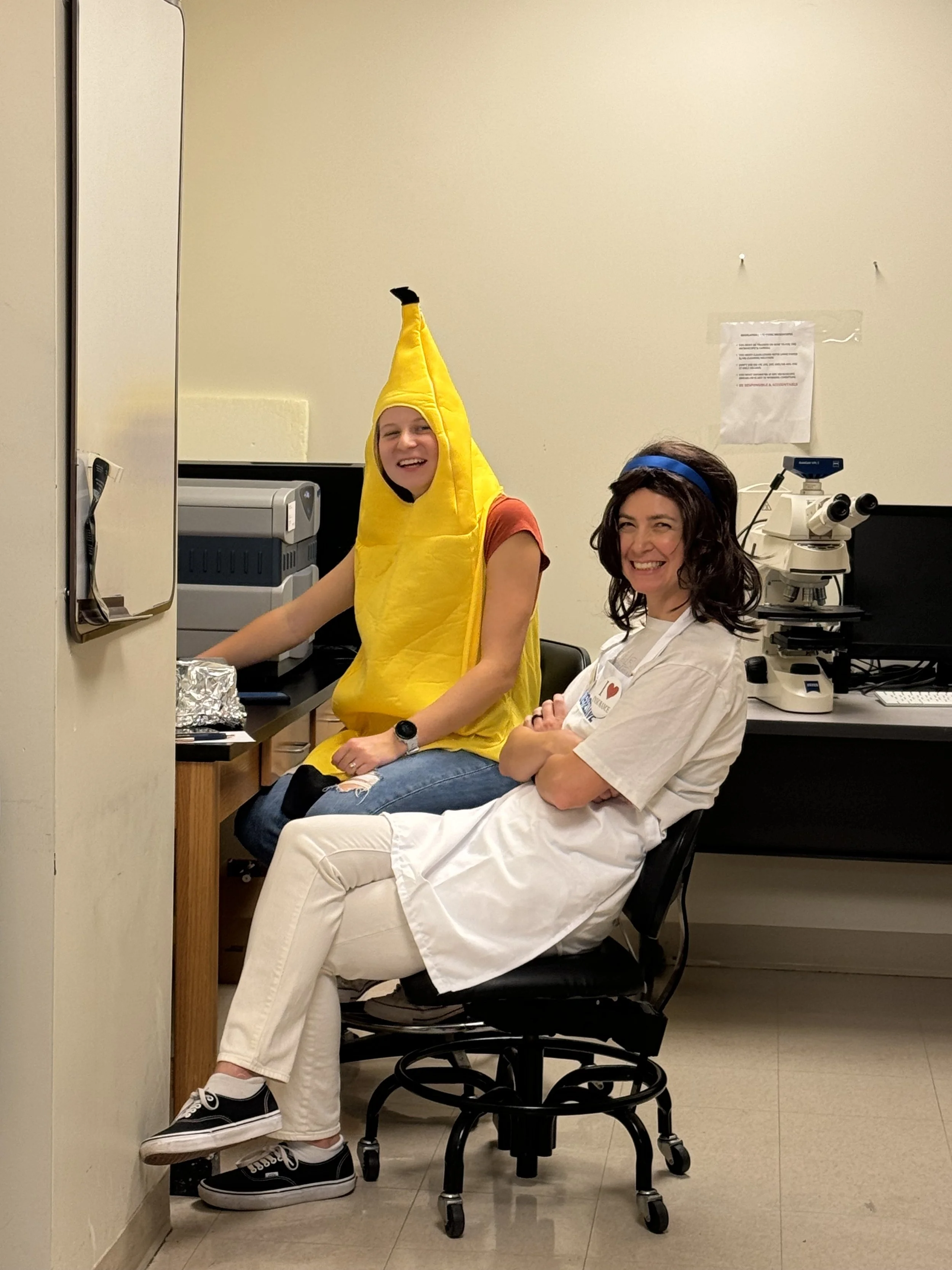 Person dressed in a banana costume sitting next to a smiling woman in medical scrubs in a laboratory or medical office.