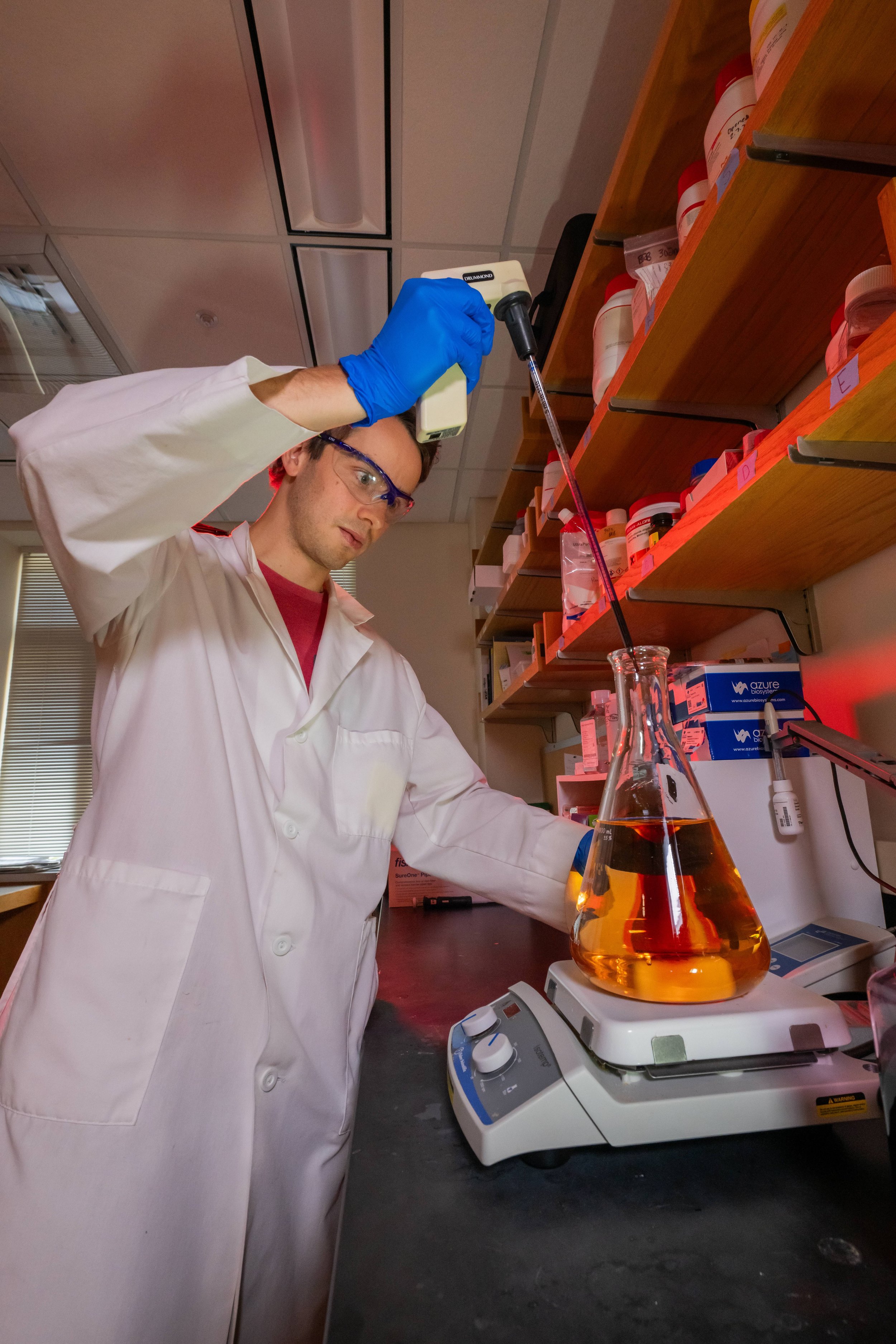 A scientist wearing a white lab coat, safety goggles, and blue gloves is pouring a liquid using a dropper into a flask with orange liquid on a digital balance in a laboratory.