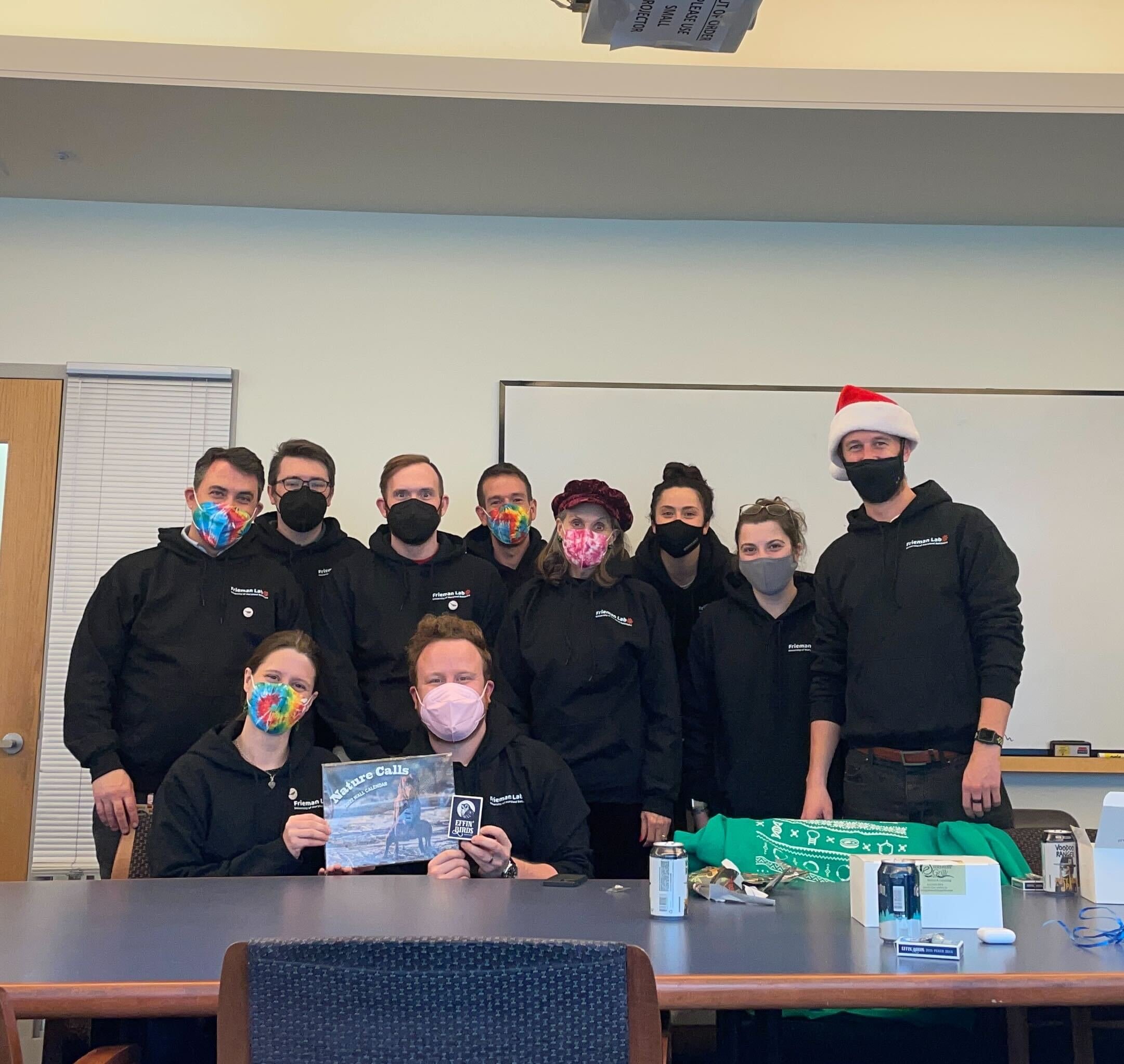 Group of ten people wearing black hoodies, some with festive masks, celebrating around a table, one person with a Santa hat, and a woman with a red Christmas hat, in a room with white walls and a dry erase board.