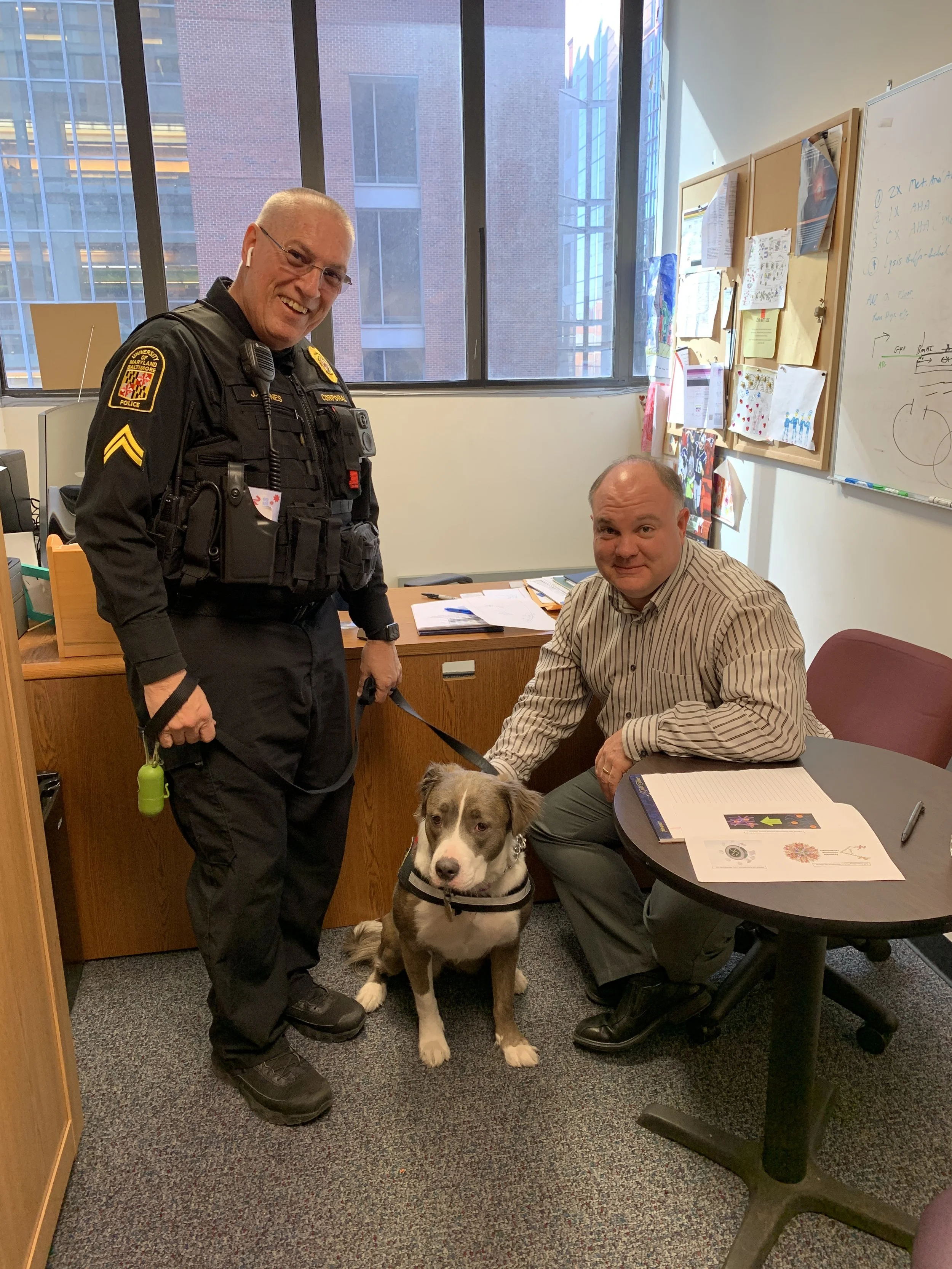 A police officer and a man sitting at a desk with a dog in an office. The police officer is standing, smiling, and wearing a black uniform. The man is seated, wearing a striped shirt, holding the leash of the dog, which is sitting on the floor lookin