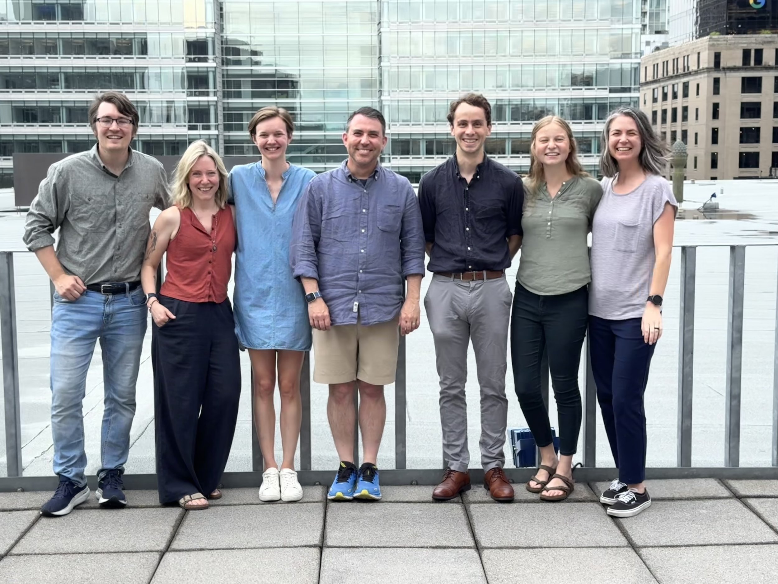 Group of seven people standing on a rooftop with a cityscape background, smiling for the camera.
