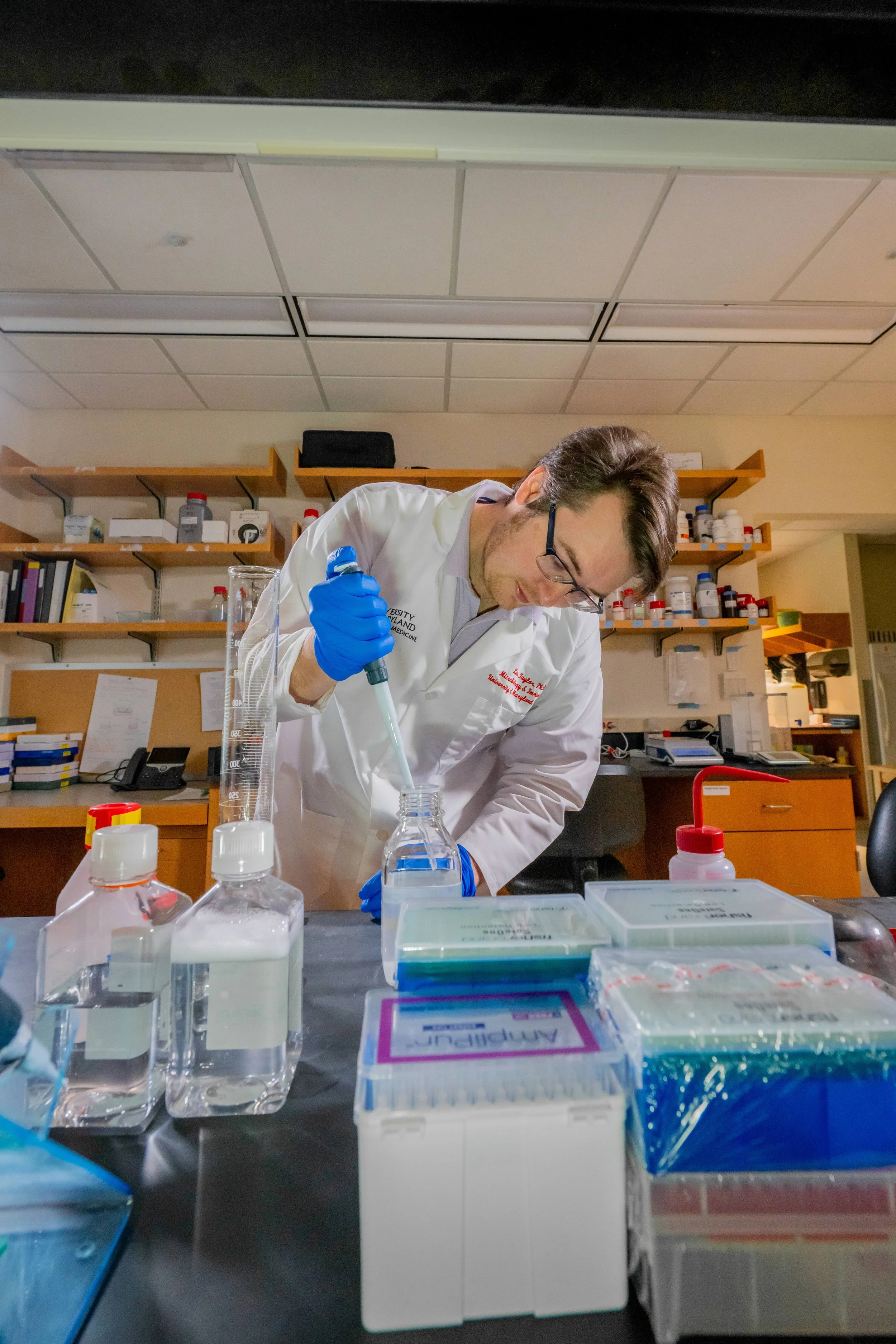 A scientist in a laboratory wearing a white lab coat and blue gloves is using a pipette to transfer liquid into a test tube. The lab bench is equipped with various bottles, containers, and scientific equipment, with shelves filled with laboratory sup