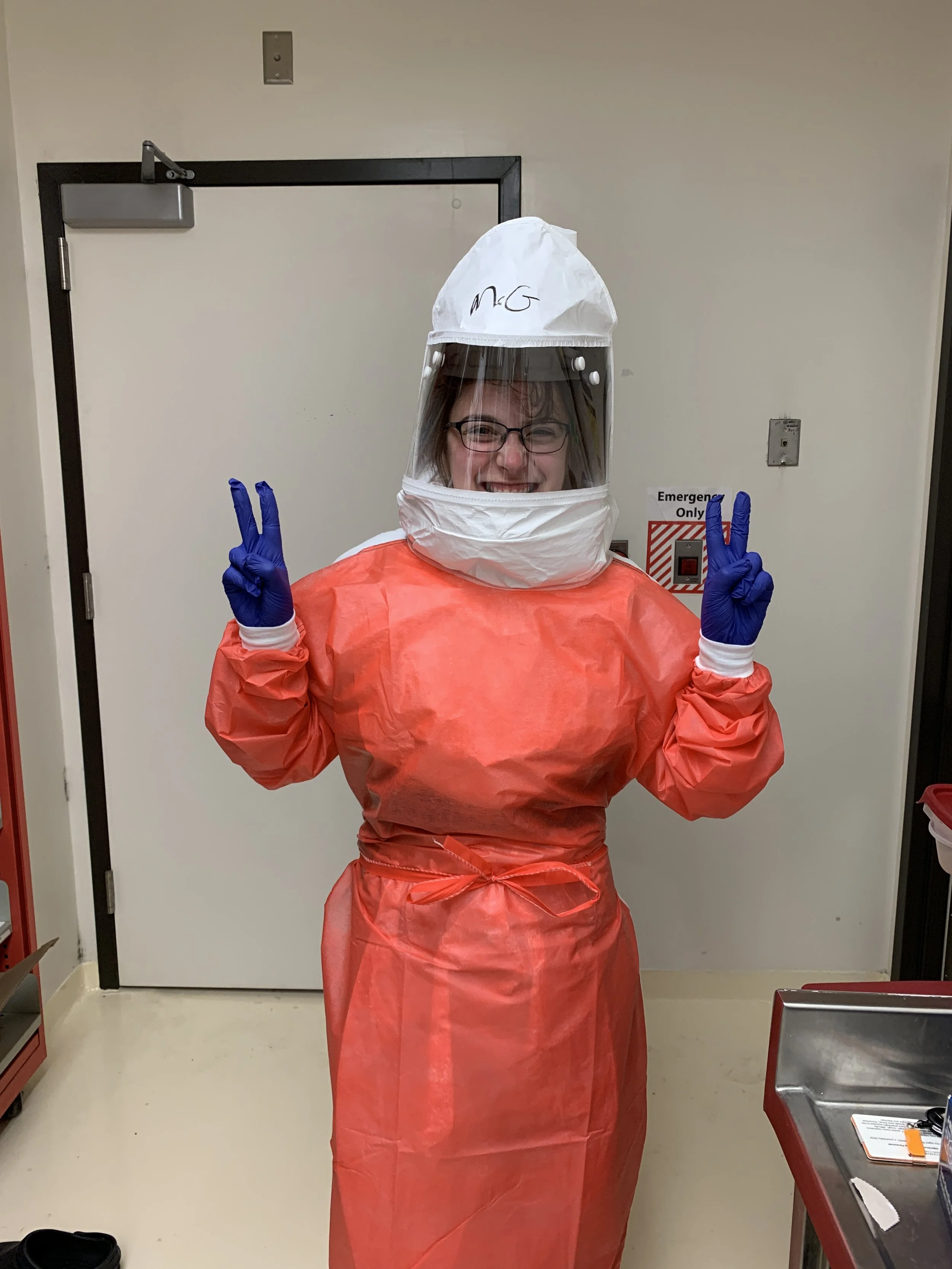 Person in full medical PPE, including a white hood with a face shield, a bright orange gown tied at the waist, and blue gloves, making peace signs with both hands in a hospital or medical setting.