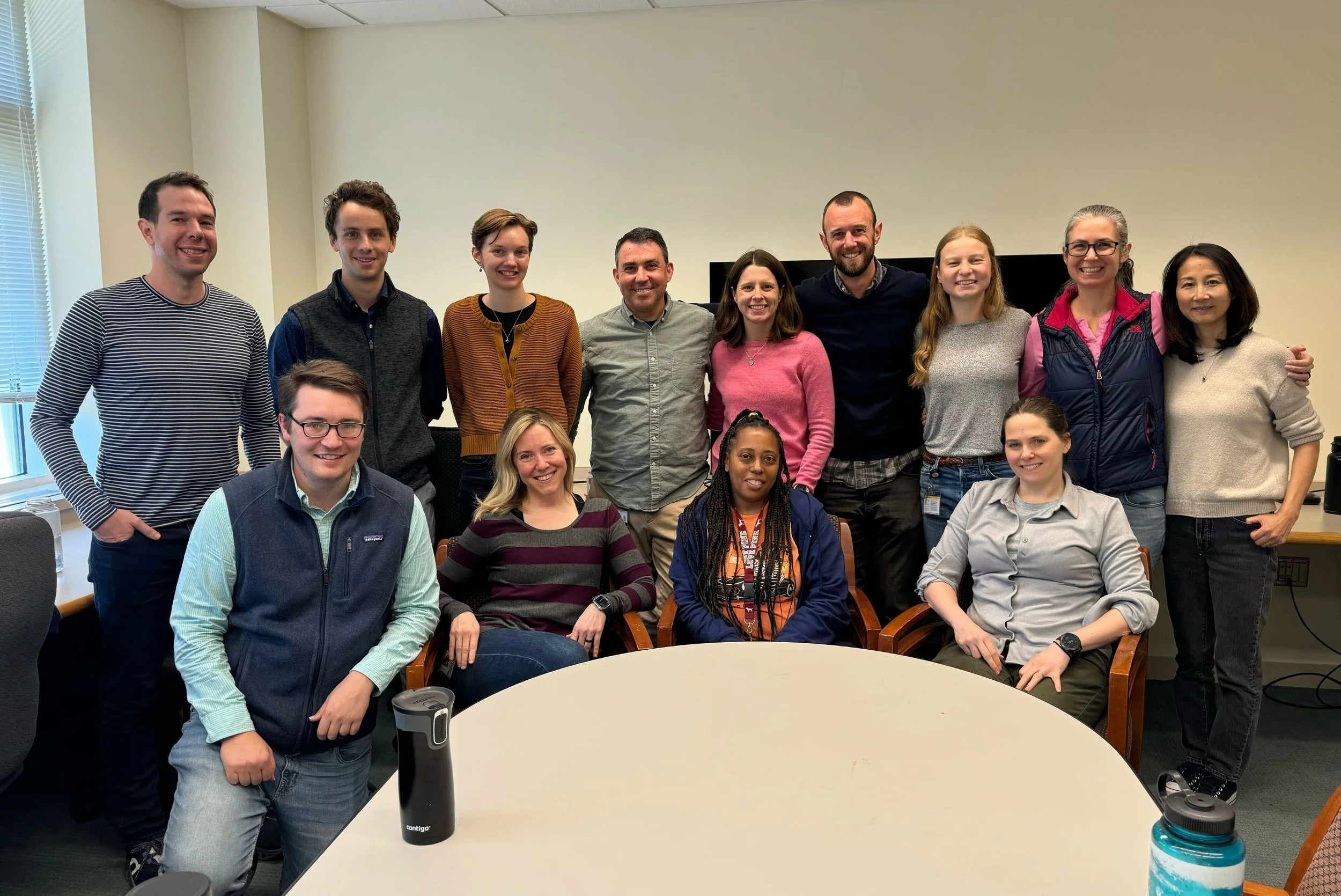 Group of 13 diverse young adults in a meeting room, some standing behind seated individuals around a round table, smiling for the camera.