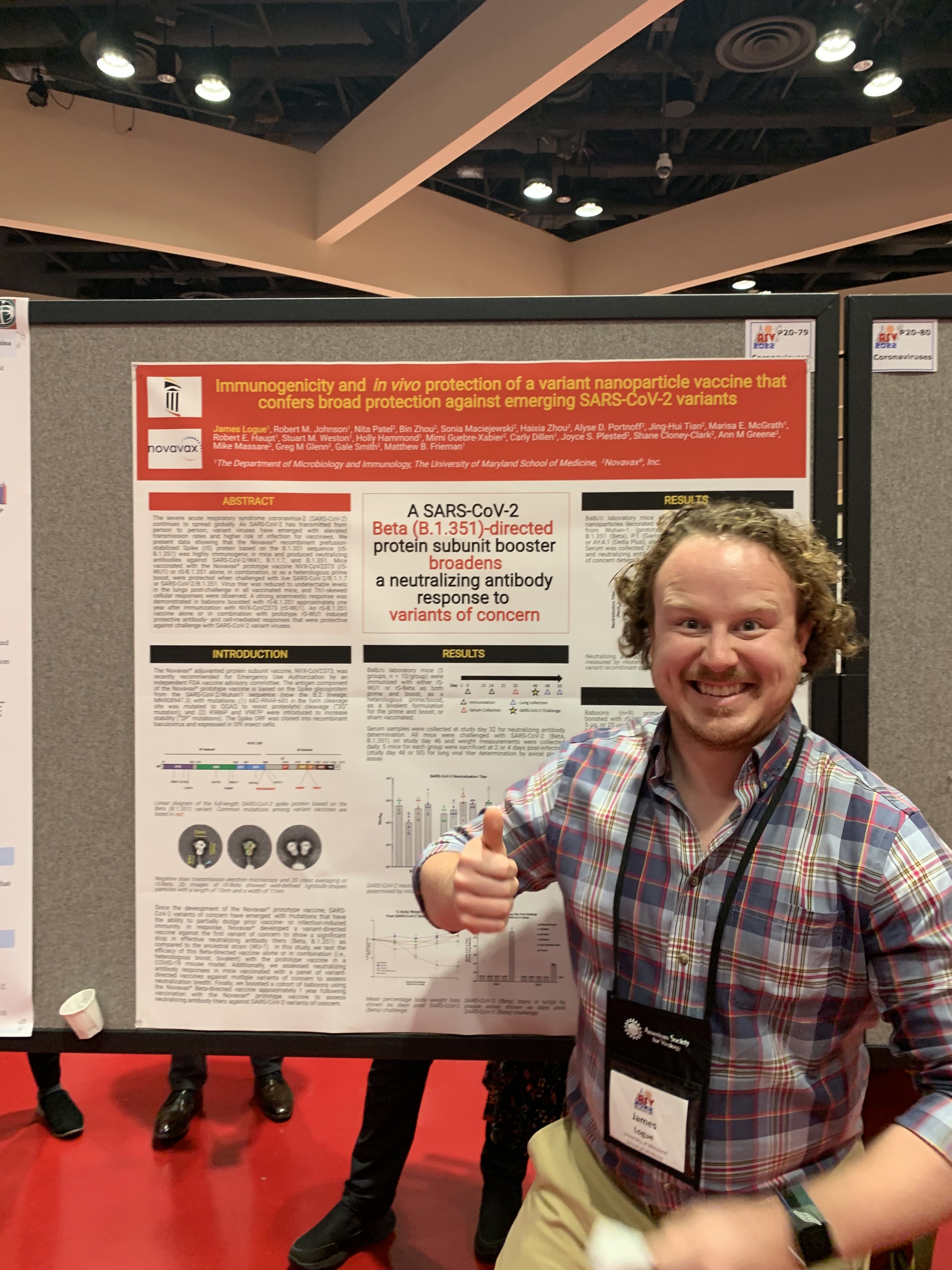 A smiling man in a plaid shirt at a scientific conference, giving a thumbs-up in front of a research poster about a SARS-CoV-2 vaccine.