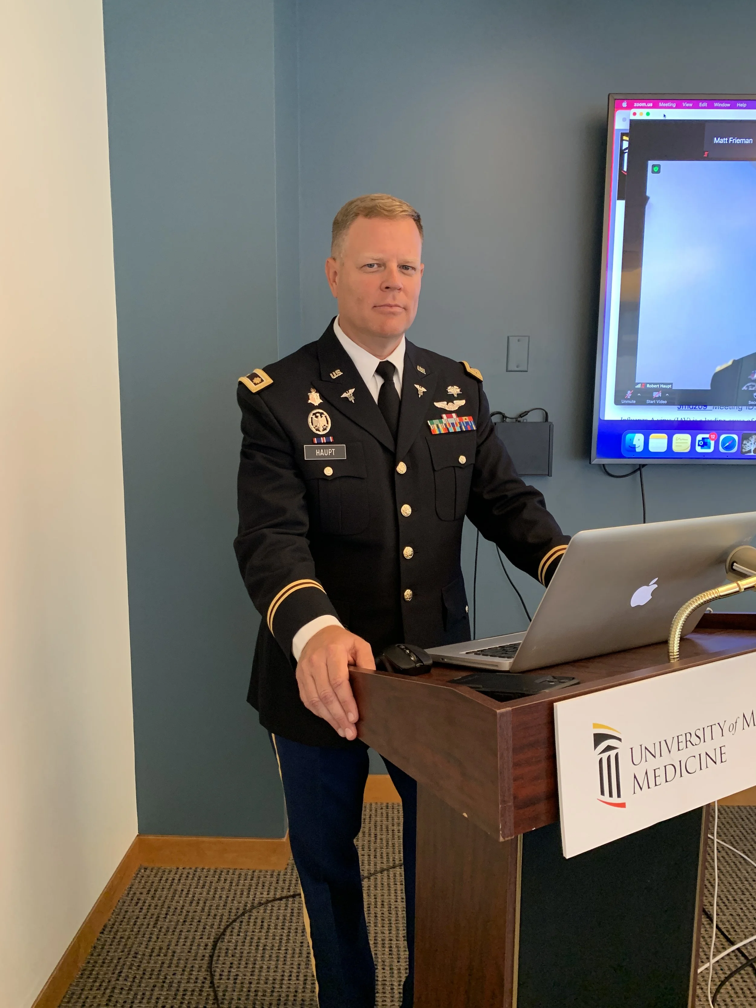 A man in a military uniform stands at a podium with a laptop, in front of a blue wall and a large monitored screen displaying a Zoom meeting. The podium has a sign that reads 'University of Medicine'.