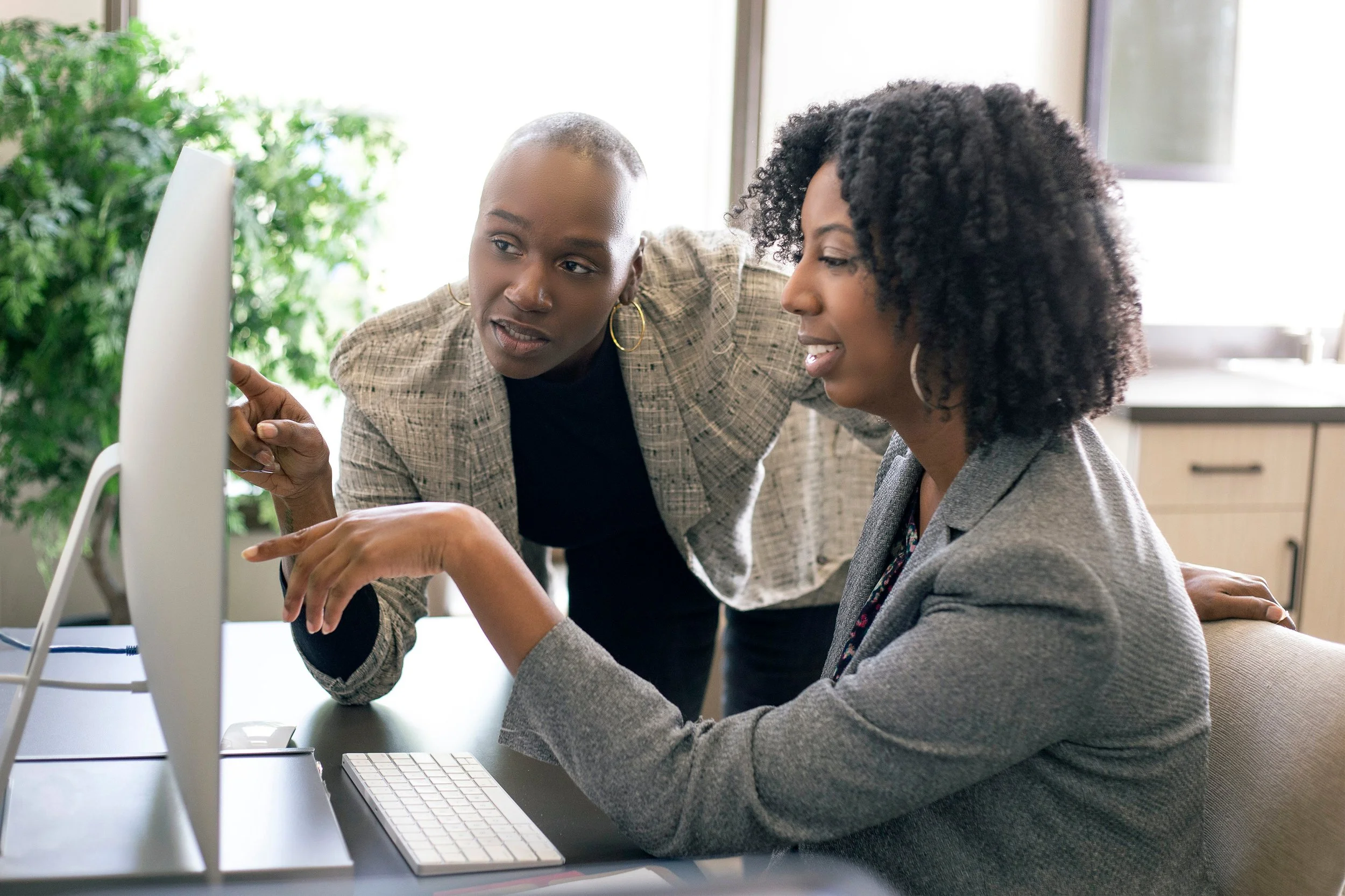 Two women working together at a computer, one pointing at the screen and explaining something to the other.