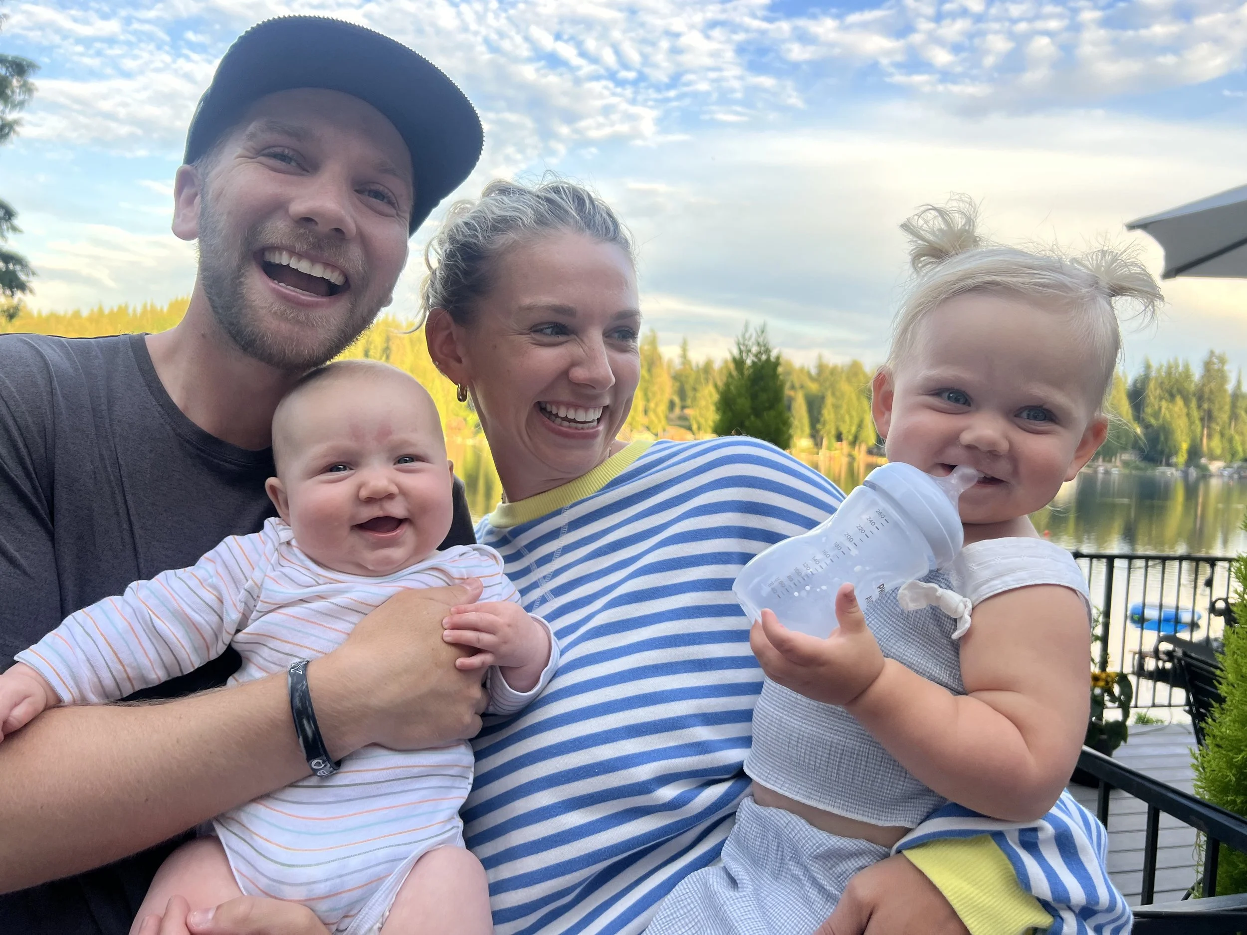 A happy family of four outdoors near a lake, including a man in a dark cap, a woman in a striped shirt, a baby in a striped onesie, and a young girl drinking from a bottle, with trees and blue sky in the background.