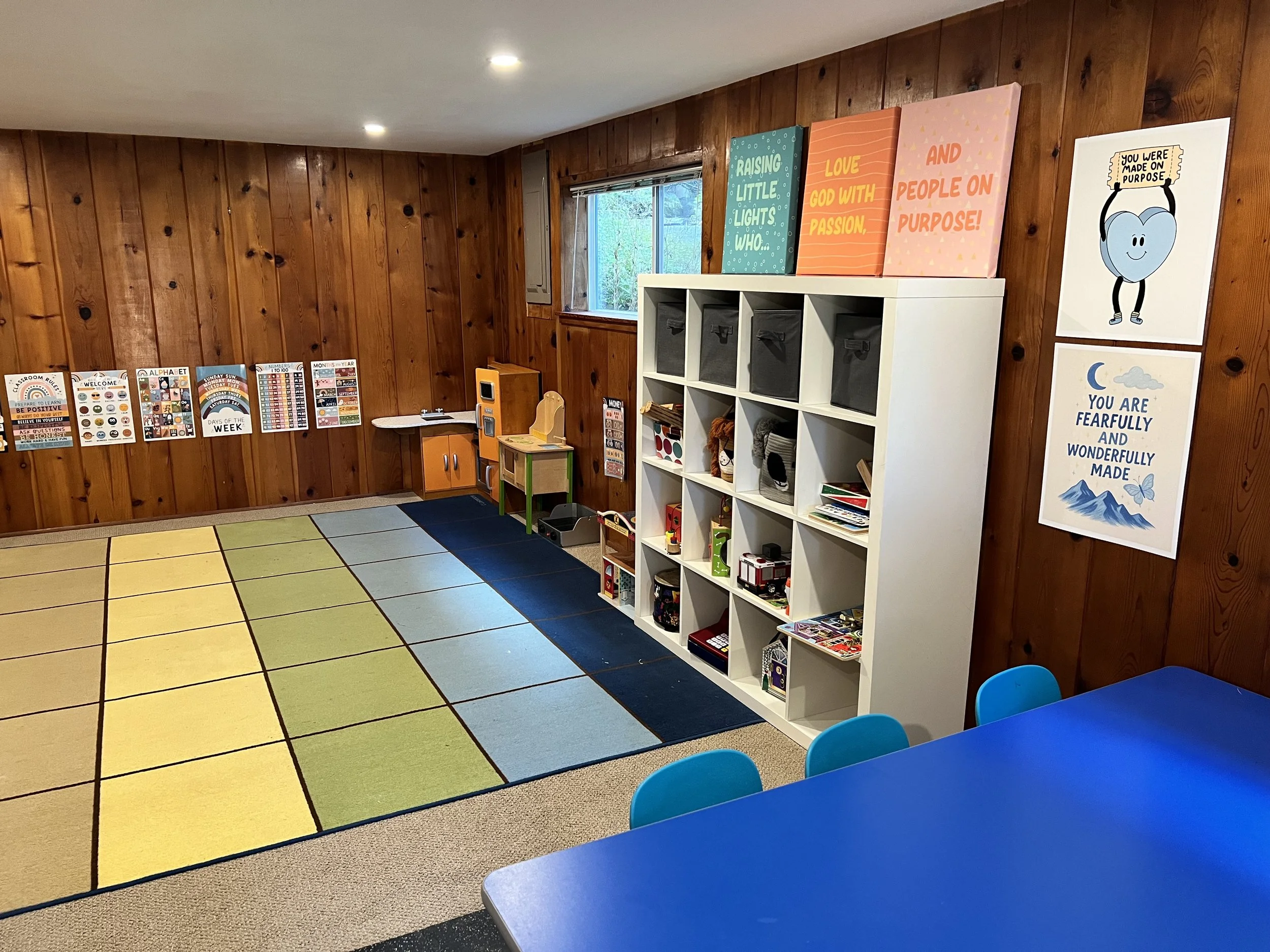 A colorful classroom with wooden walls, a window, and a multi-colored rug. There are posters on the wall, a white cubby shelf with bins, and a blue table with blue chairs.