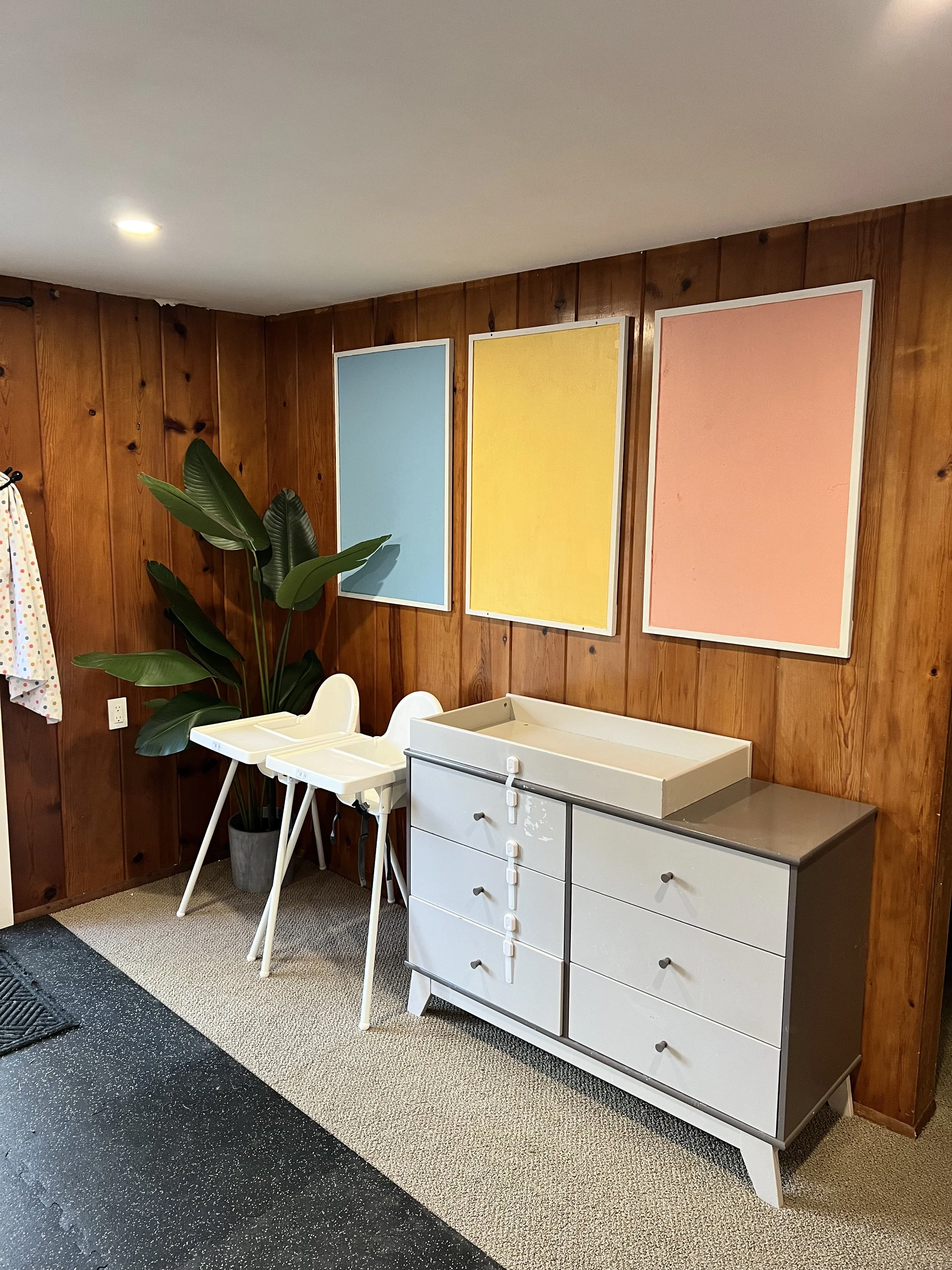 Interior of a room with three colorful abstract paintings on a wood-paneled wall, a potted plant, two white high chairs, and a white and gray changing table.