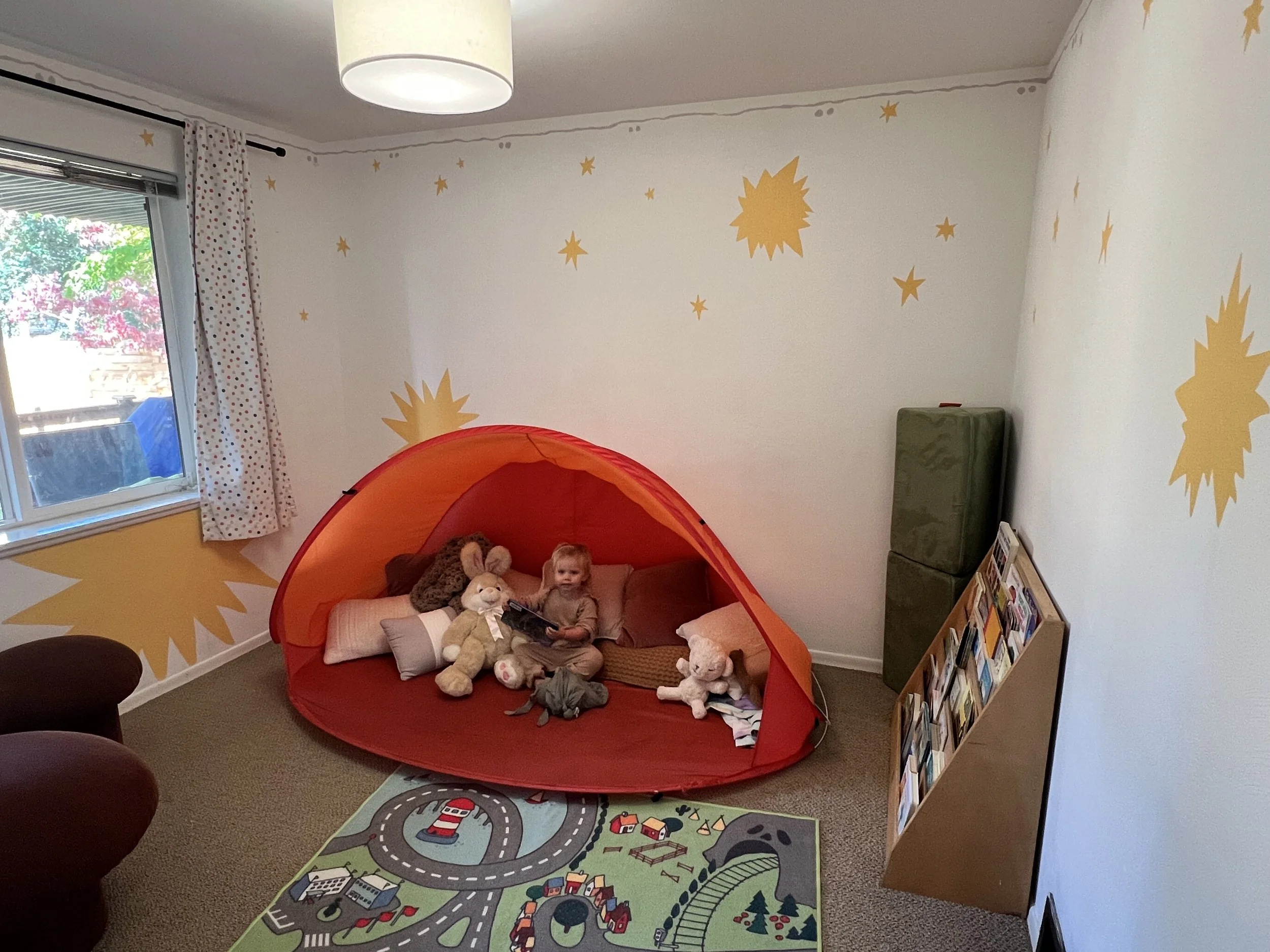 A children's playroom with a small orange tent, stuffed animals, and a young girl sitting inside reading. The room has yellow star and sun decals on white walls, a window with polka-dot curtains, and a colorful play mat on the carpeted floor.