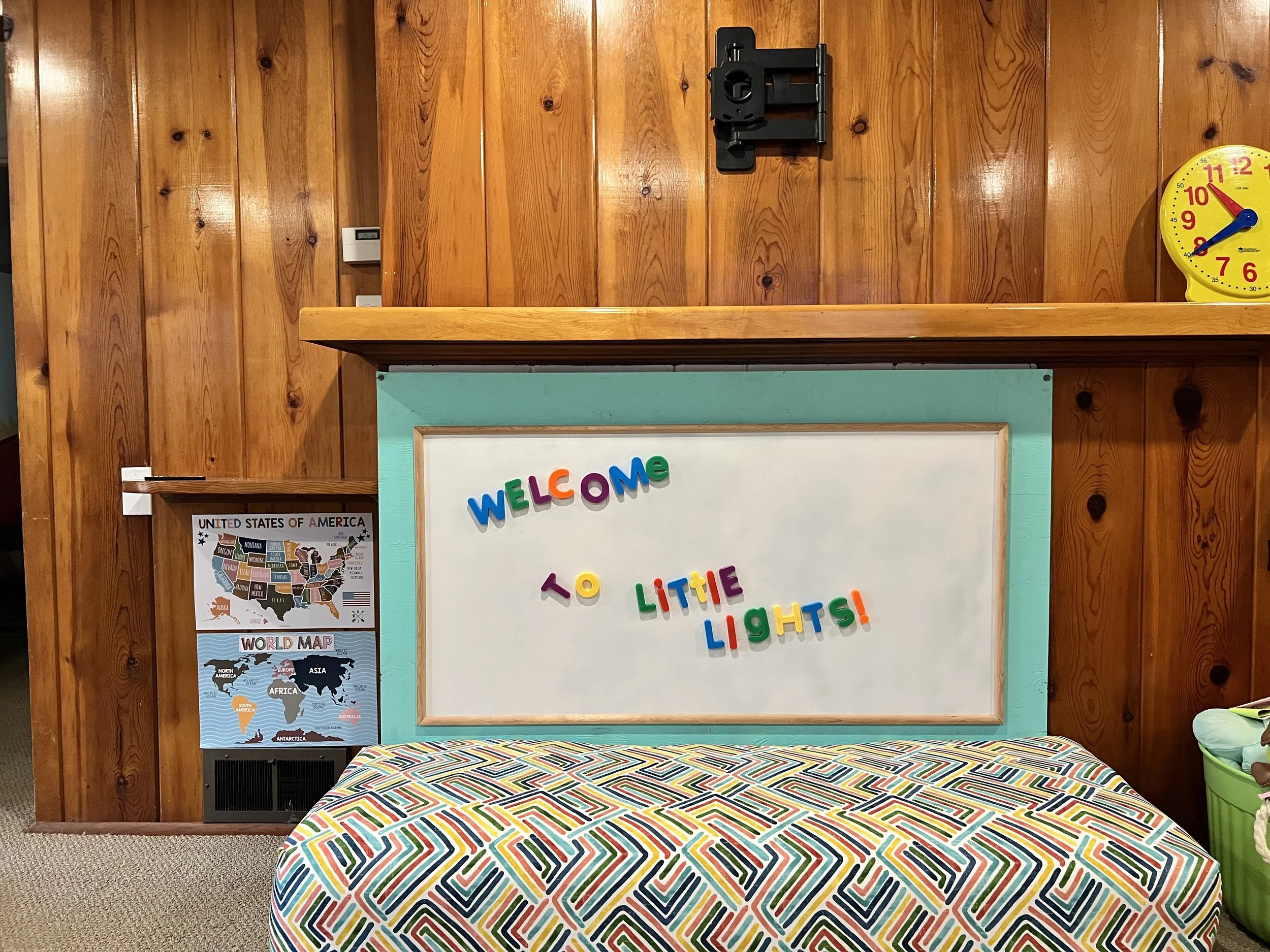 A classroom or child’s activity area with a wooden wall background. There is a colorful, patterned tablecloth on a table in front. A whiteboard displays multicolored magnetic letters spelling "Welcome to Little Lights!" To the left, there are educational posters including a United States map and a world map. To the right, there is a yellow clock and a black wall-mounted holder. The wall and furniture have a warm, inviting atmosphere.