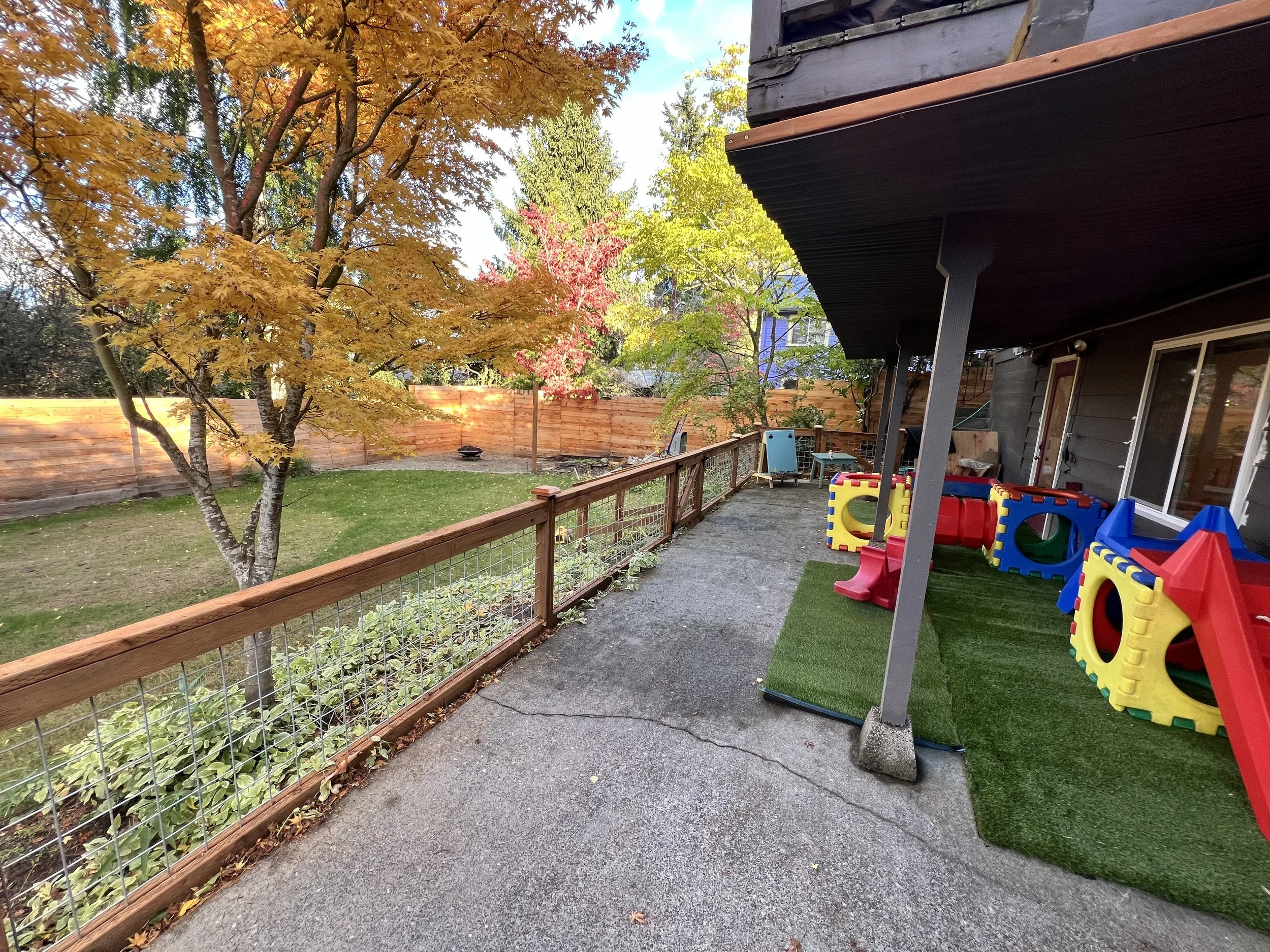 A backyard patio with a grassy yard, colorful trees with autumn leaves, a wooden fence, and children's play structures including slides and tunnels covered with artificial grass.