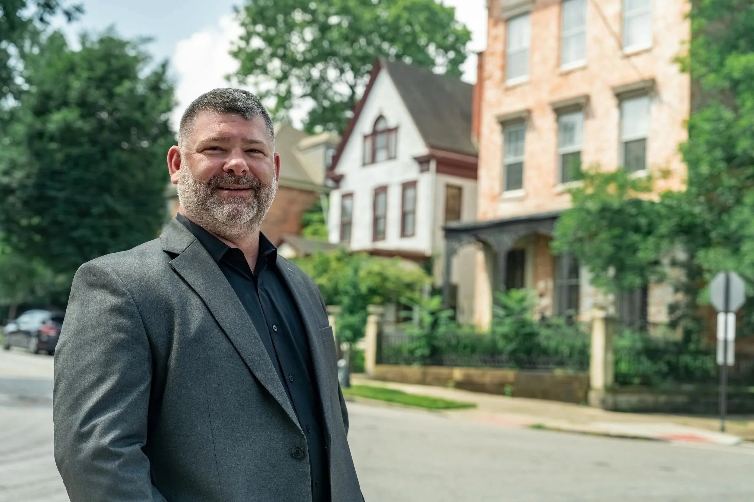 A man in a dark suit standing outdoors in front of old brick and wood residential buildings surrounded by trees, smiling at the camera.