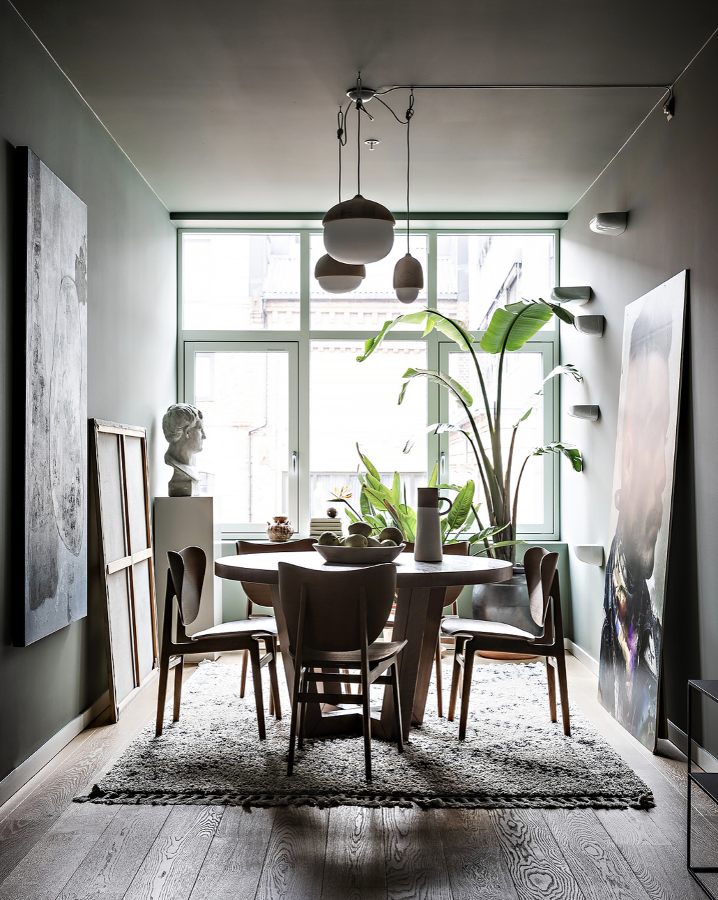 Modern dining room with a round wooden table, six chairs, large window with green framing, indoor plants, artwork, and decorative objects on the table and windowsill.