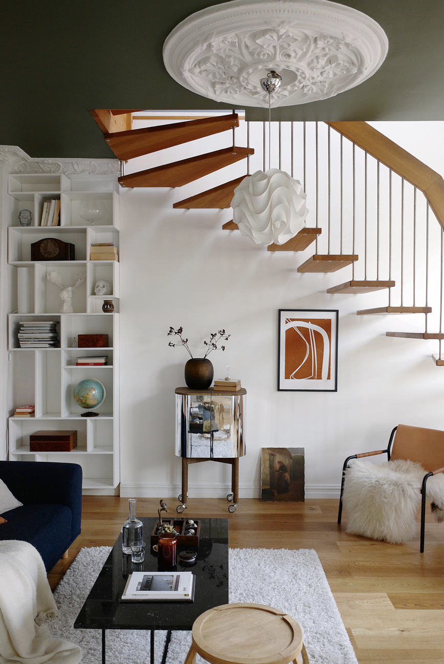Living room with a modern wooden staircase, white shelving unit, decorative artwork, a black coffee table with magazines and candles, and an orange chair with a sheepskin throw. Ceiling has an ornate white medallion and a sculptural pendant light.