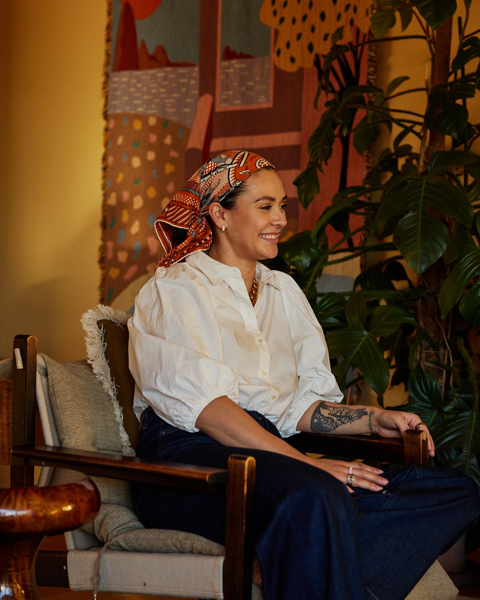 A woman sitting on a chair, smiling, wearing a white blouse, dark jeans, and a colorful headscarf. She has tattoos on her right forearm and is in a cozy, art-filled room with a large leafy plant behind her.